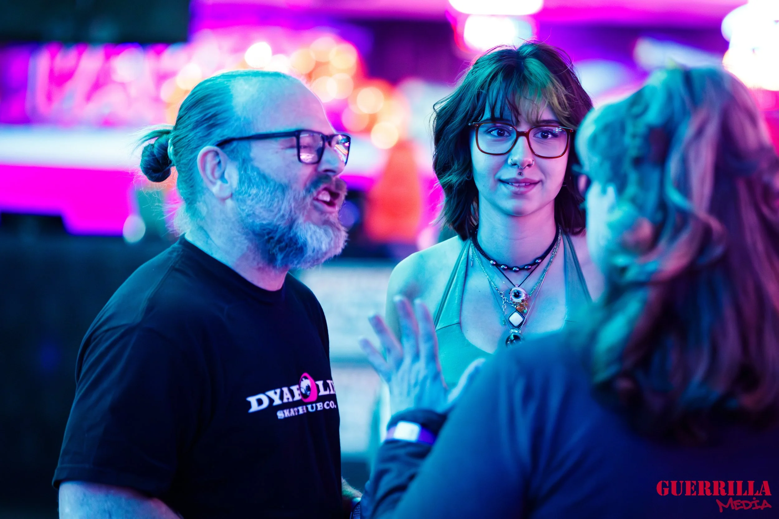 Three people engaged in conversation at a neon-lit skate park. The man on the left has a long gray beard, glasses, and a man bun, wearing a black T-shirt that reads 'DYAO' and features a logo. The woman in the middle has shoulder-length hair, glasses
