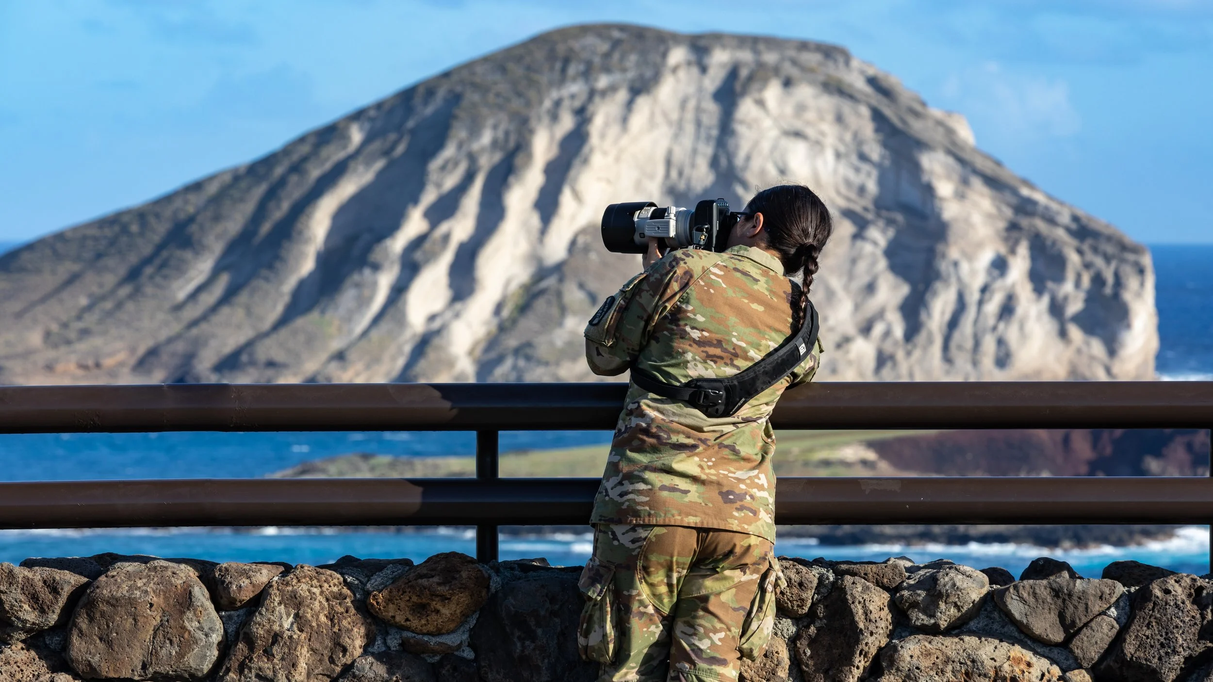 A person dressed in camouflage military uniform taking a photograph with a camera, standing in front of a rocky stone wall and overlooking a large island with some greenery and cliffs by the ocean.