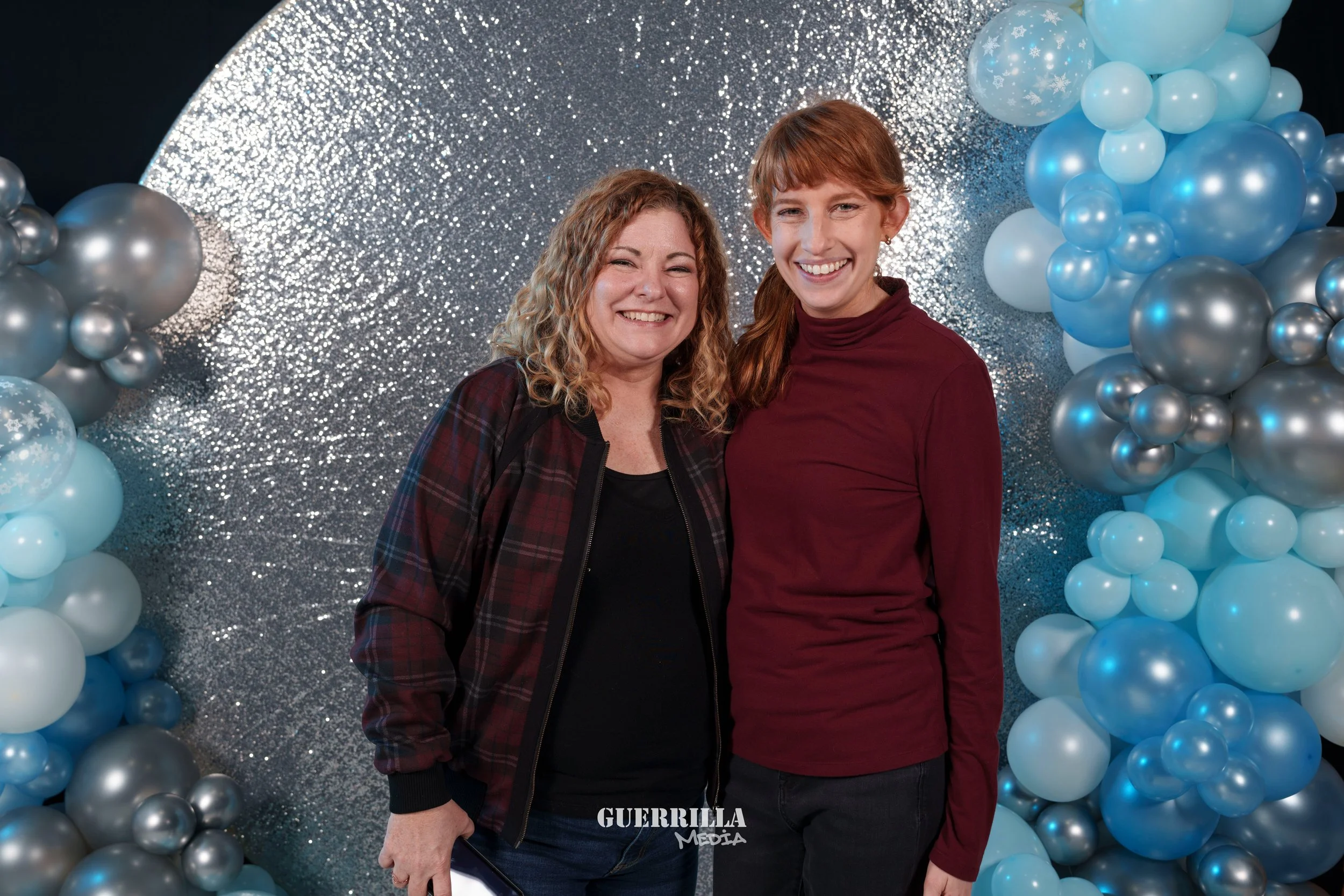 Two women smiling and standing close together in front of a festive backdrop with silver and blue balloons and a sparkling silver curtain.