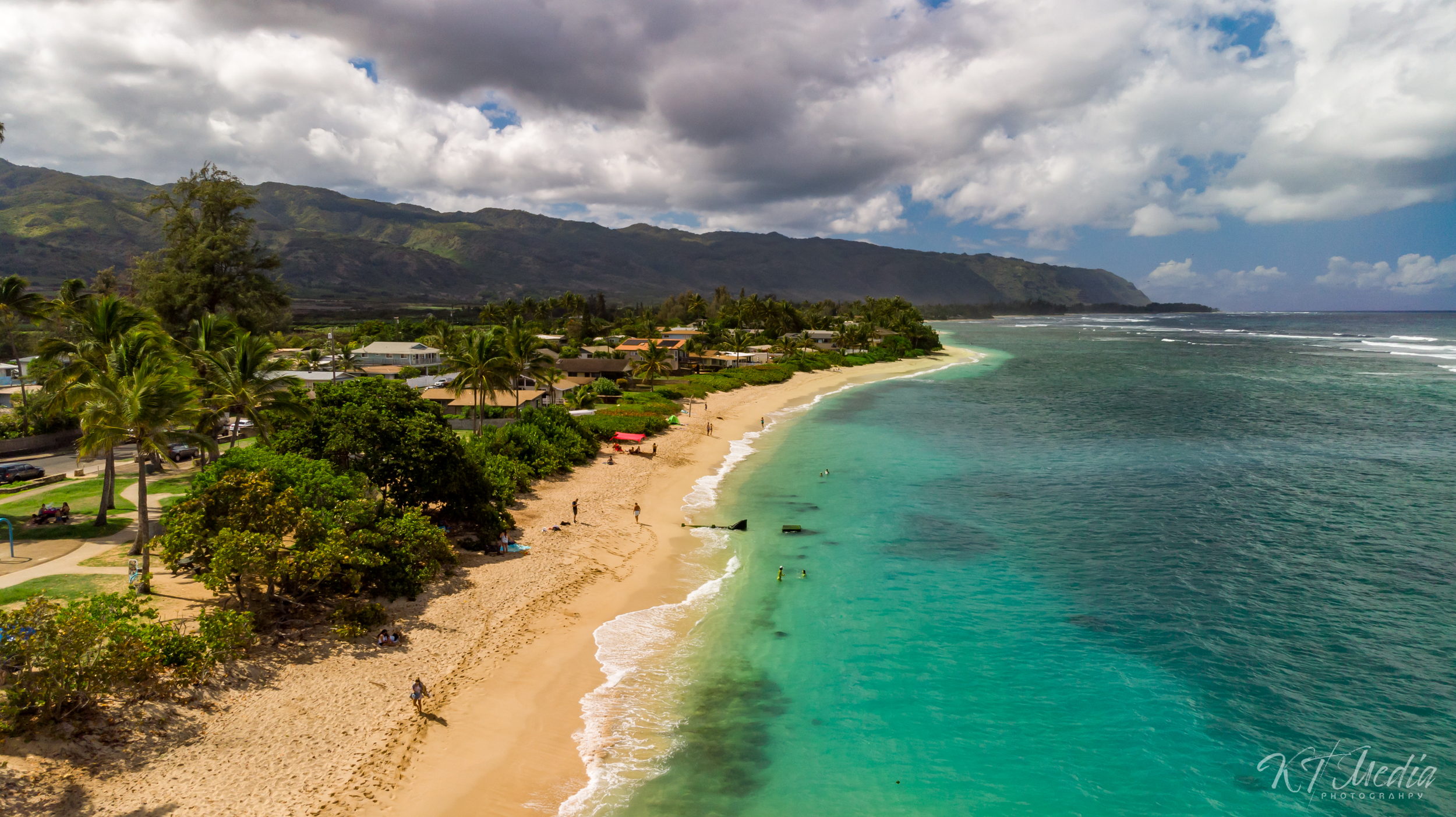 A coastal view showing a sandy beach with palm trees, houses, and lush greenery in front of mountains under a partly cloudy sky, with people swimming and walking along the shoreline.
