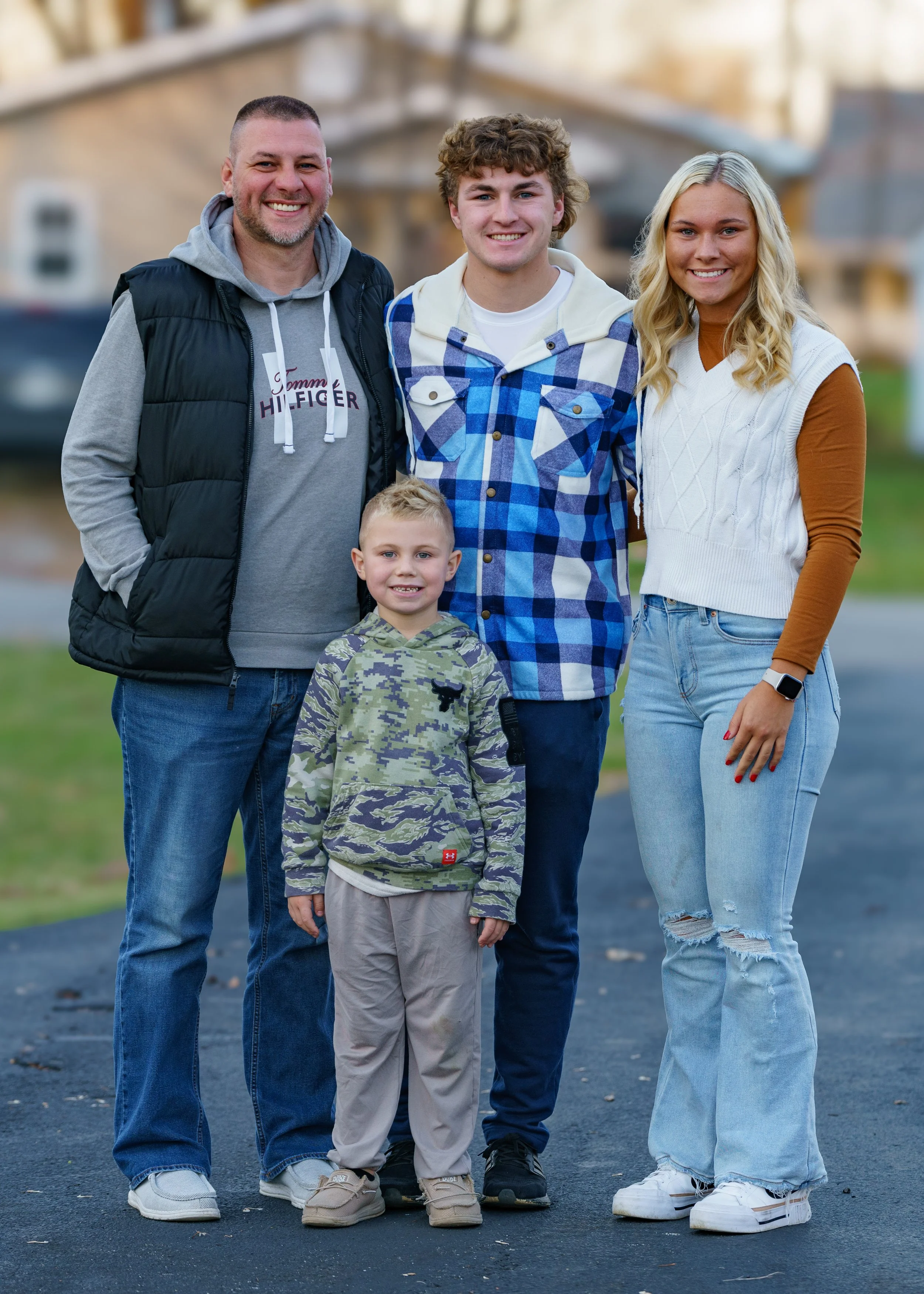 A group of five people, a man, a woman, and three boys, standing outdoors on a residential street, smiling at the camera. The man is wearing a gray hoodie and black vest, the woman is in a white sweater vest and jeans, the older boy is in a blue chec