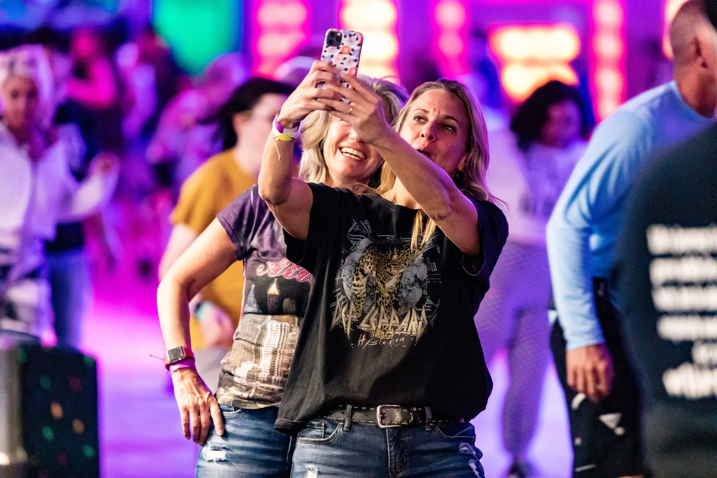 Two women taking a selfie together at a lively indoor event with colorful lights and other people in the background.