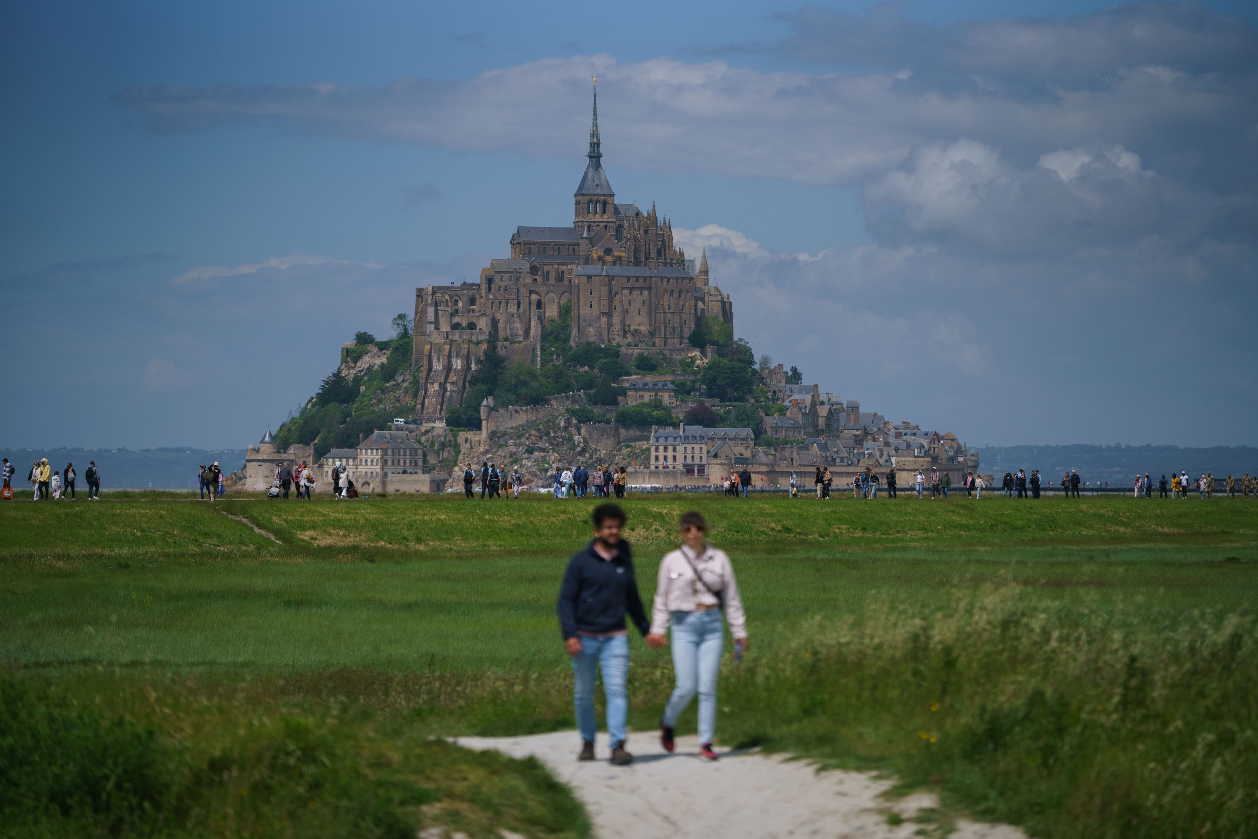 The image shows Mont Saint-Michel, a historic island commune in France, with a large, medieval-style abbey and village on top of a rocky hill. In the foreground, a young couple is walking hand-in-hand on a grassy path, while many other people are sca
