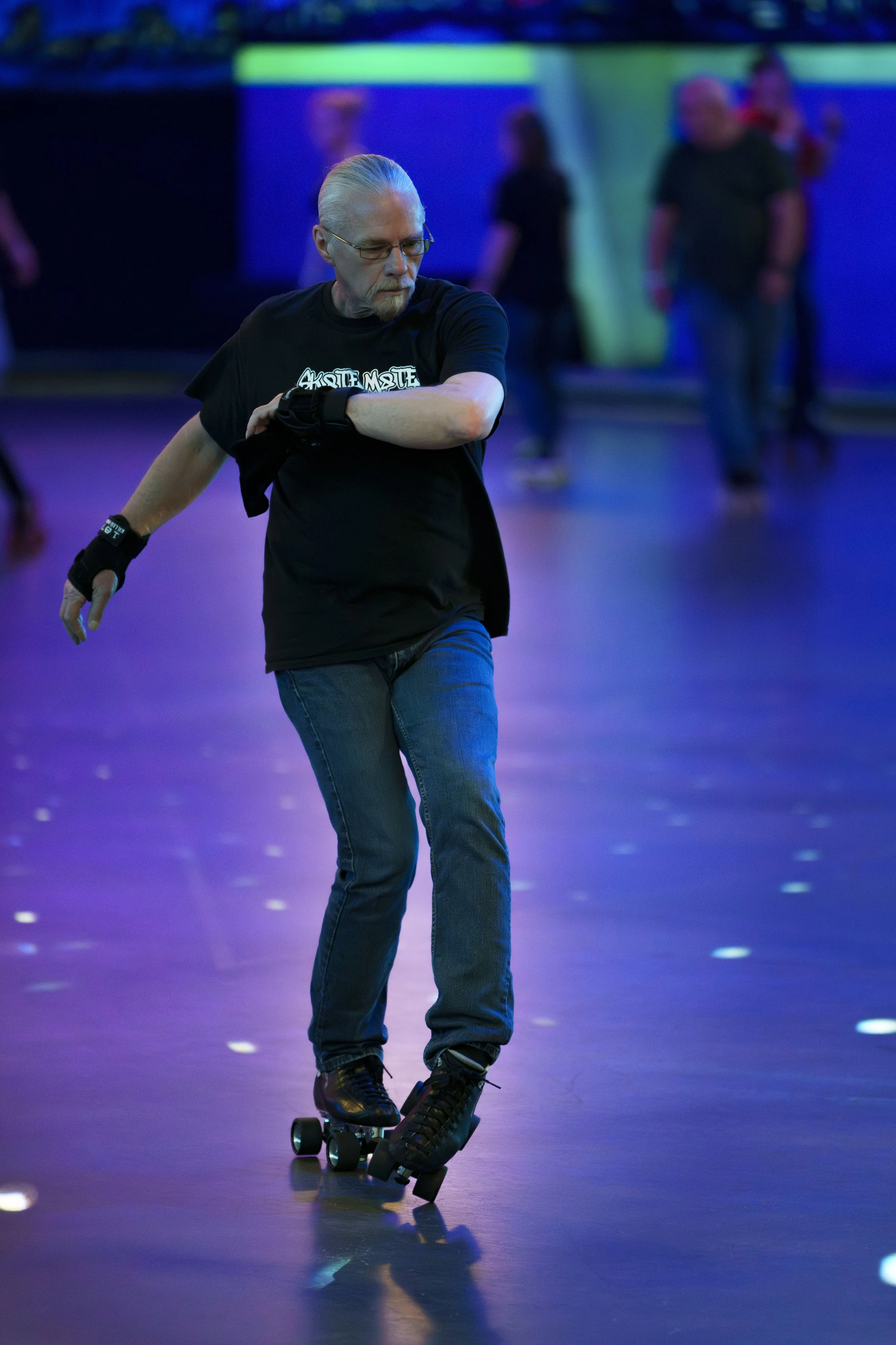 A man with gray hair and glasses roller skating inside a rink with neon lighting.
