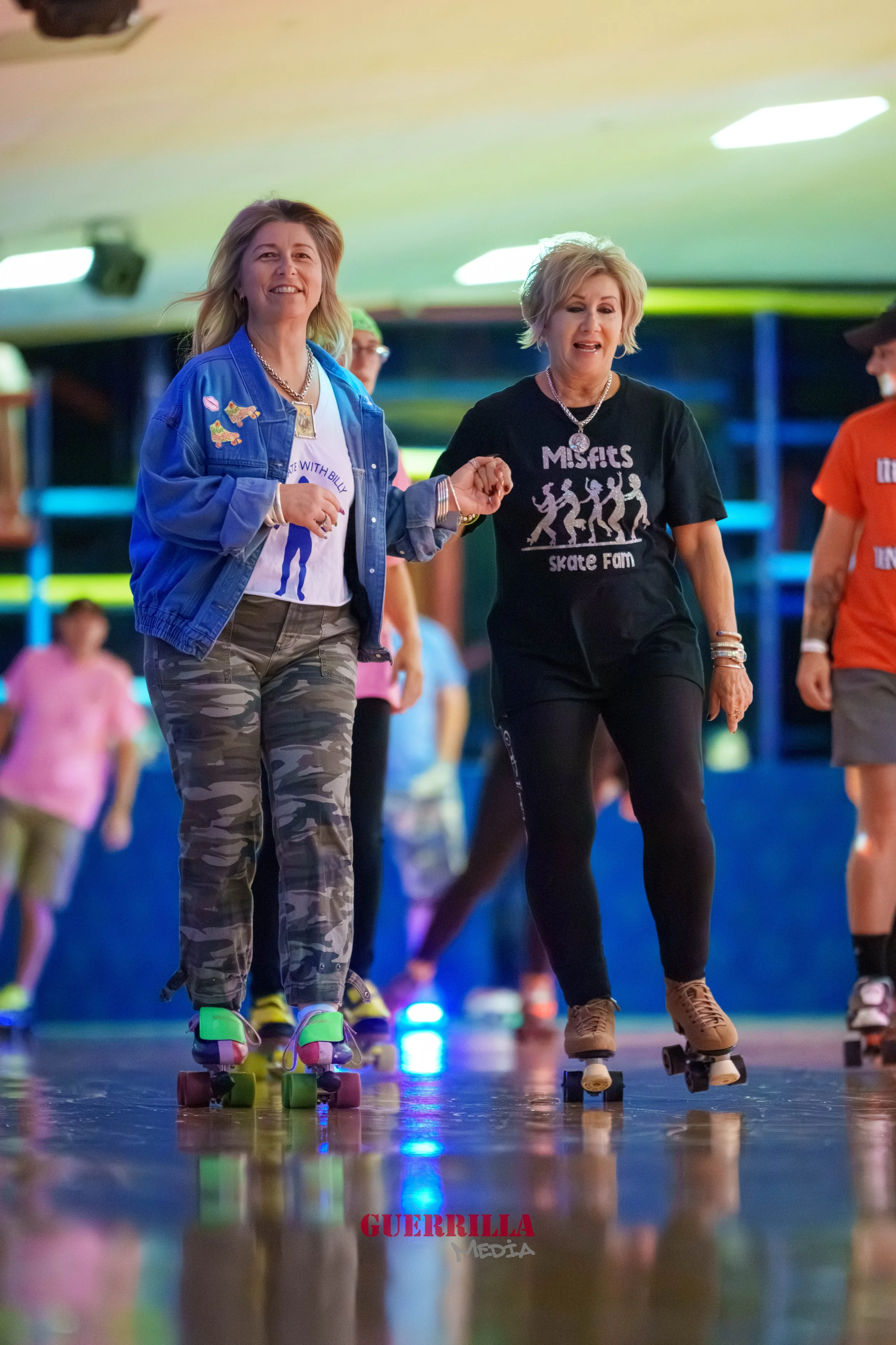 Two women roller skating indoors, holding hands and smiling, surrounded by other skaters.