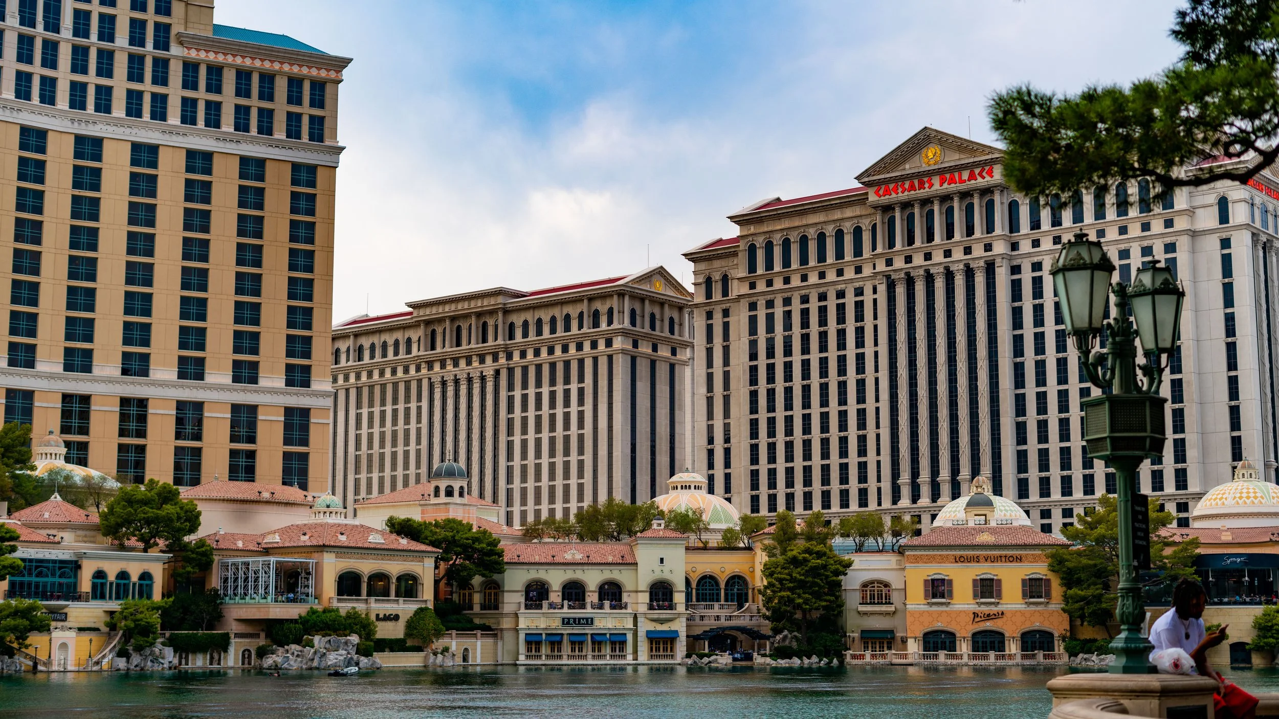 View of Caesars Palace hotel and casino in Las Vegas, Nevada, with the lake and promenade in the foreground, and a person sitting on the edge of the promenade near a green street lamp.