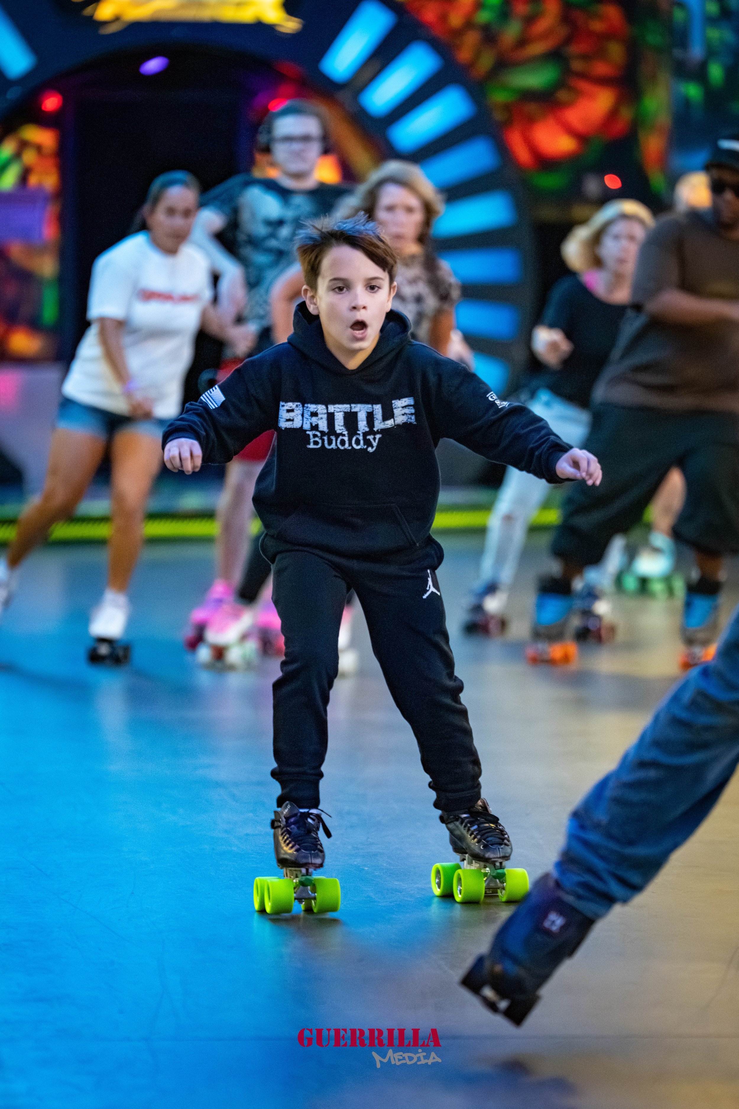 A boy in a black hoodie and pants roller skating indoors, with a surprised expression on his face, while a group of people roller skate behind him in a colorful, neon-lit arcade or roller skating rink.
