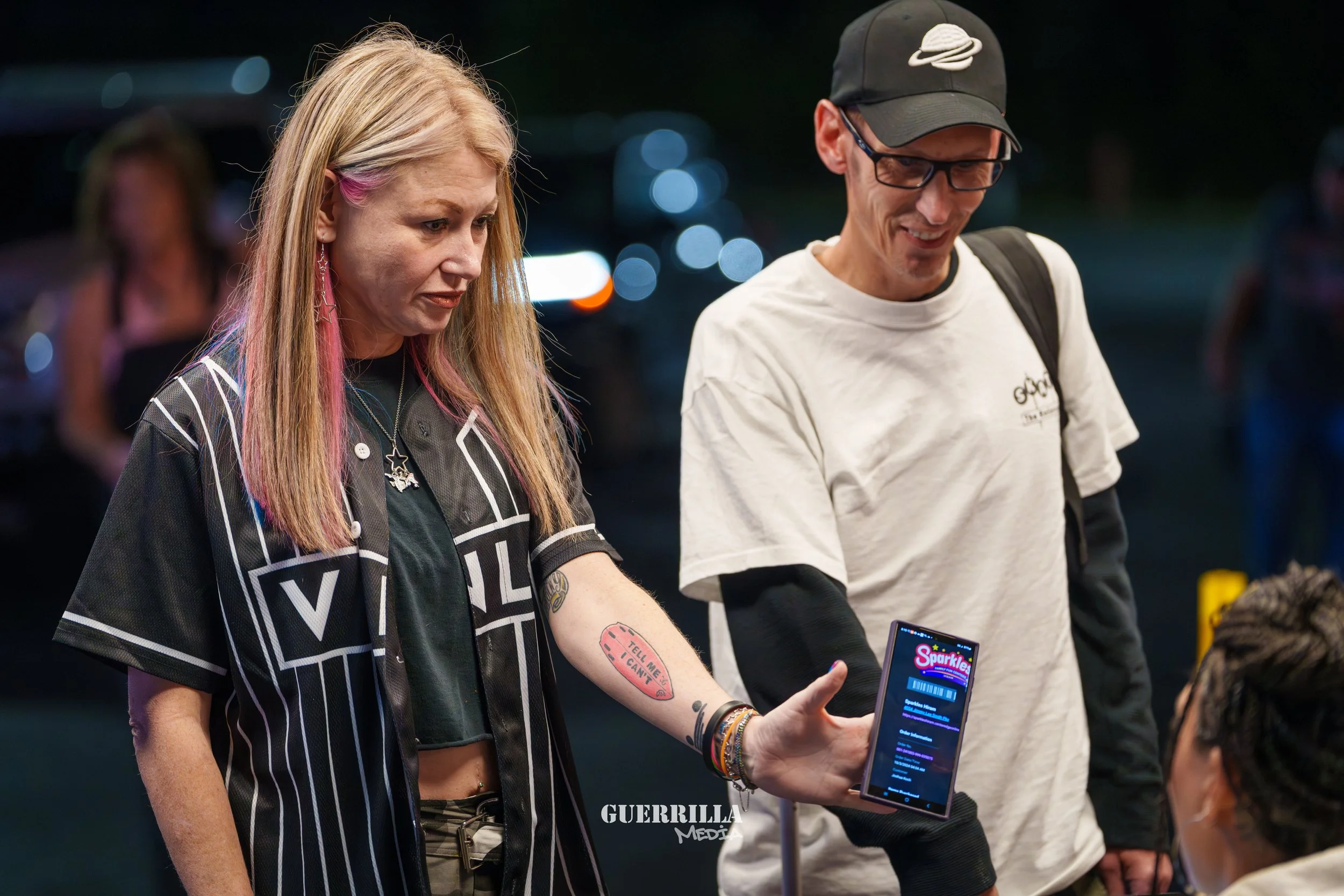 A woman with blonde hair and pink streaks showing her phone to a young man. The woman is wearing a black and white striped shirt, multiple bracelets, and has tattoos and jewelry. The young man is wearing glasses, a white T-shirt, and a black cap with