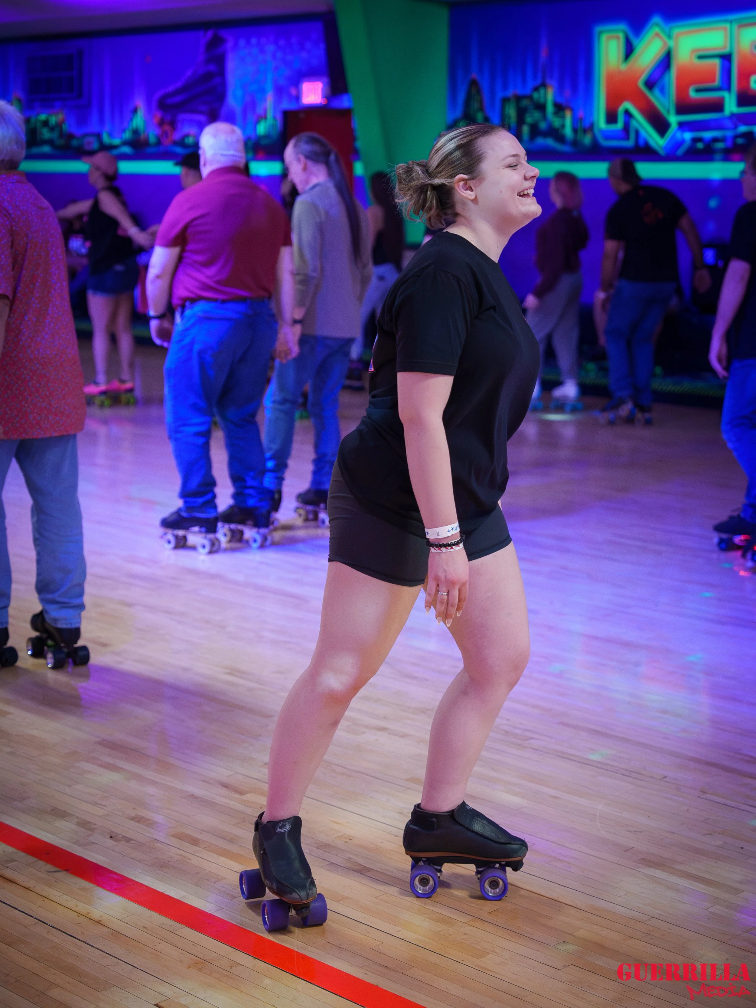 A young woman roller skating and smiling in an indoor roller skating rink with neon lighting and other skaters in the background.
