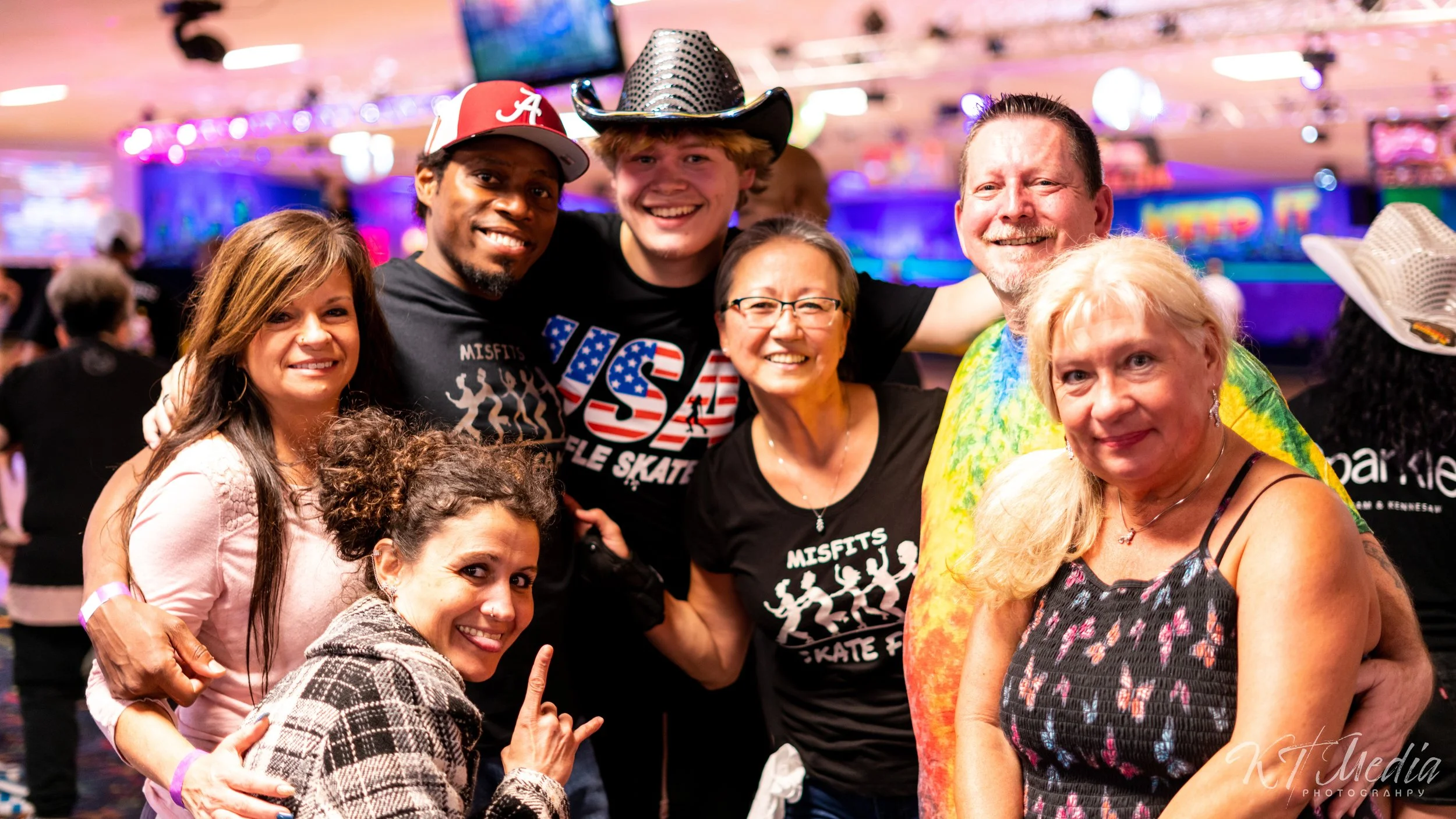 A group of seven diverse people smiling and posing together at a lively event with colorful lights in the background. They are wearing casual and themed attire, including T-shirts and a cowboy hat, and seem to be enjoying a fun atmosphere.