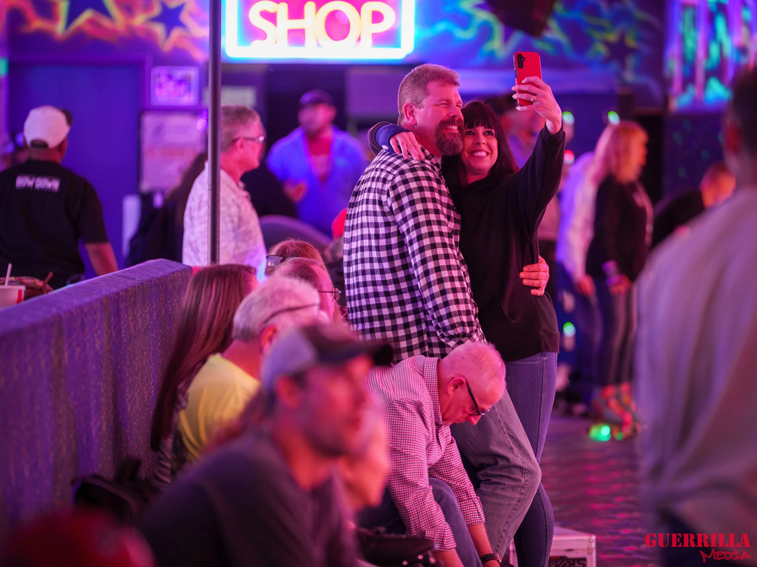 A man and woman take a selfie together at an indoor entertainment venue with colorful neon lights. The man has a beard and wearing a checkered shirt, the woman has dark hair and is smiling. Other people are seated and standing around, some watching, 