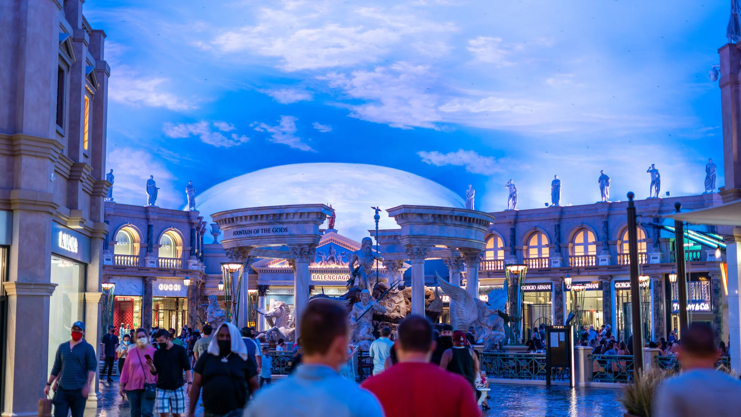 People walking in a shopping plaza with classical sculptures and a fountain, under a blue sky with clouds