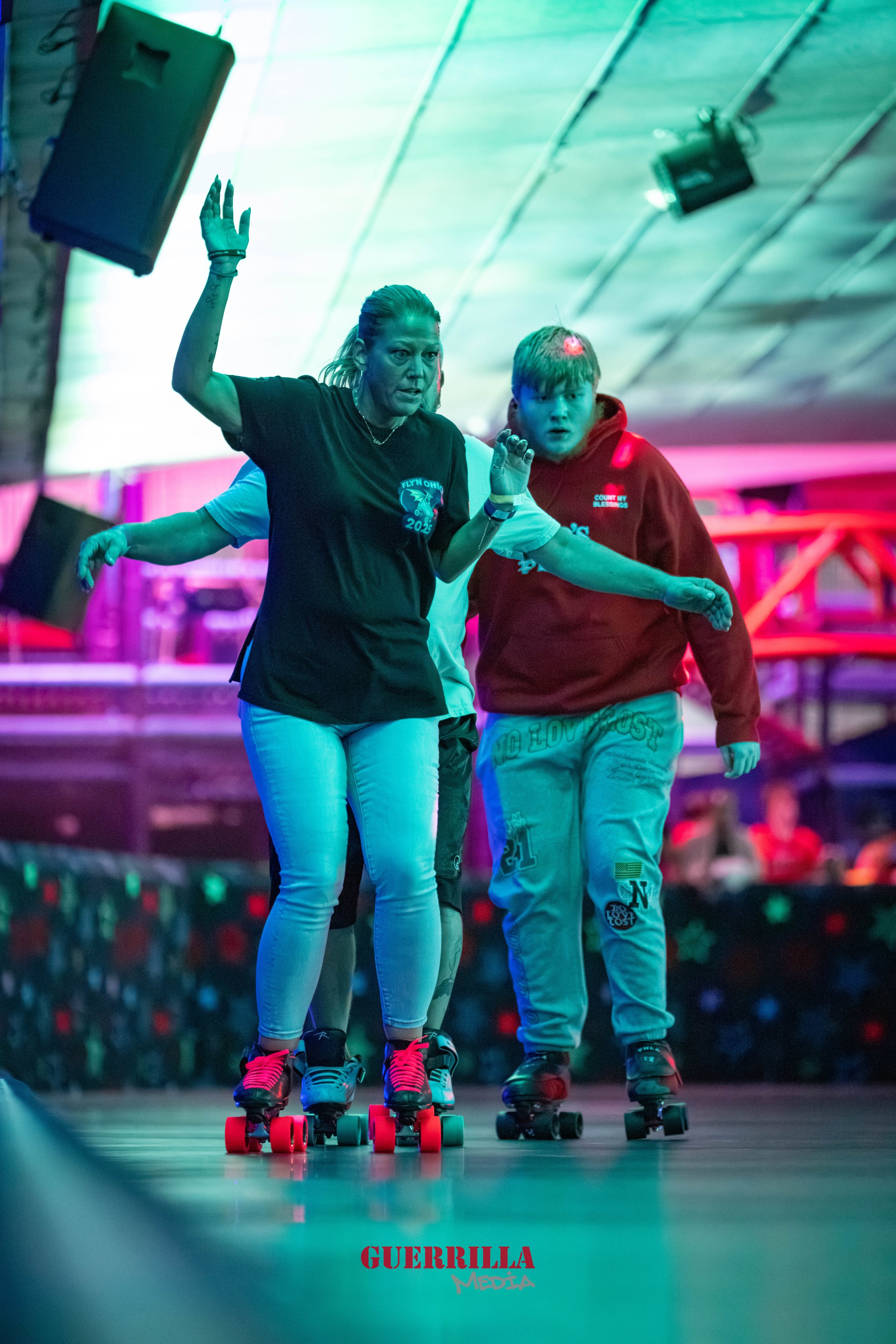 People roller skating indoors under colorful, neon lighting with a woman guiding a young boy. The background is blurred and there is a red logo at the bottom that reads 'GUERRILLA MEDIA.'