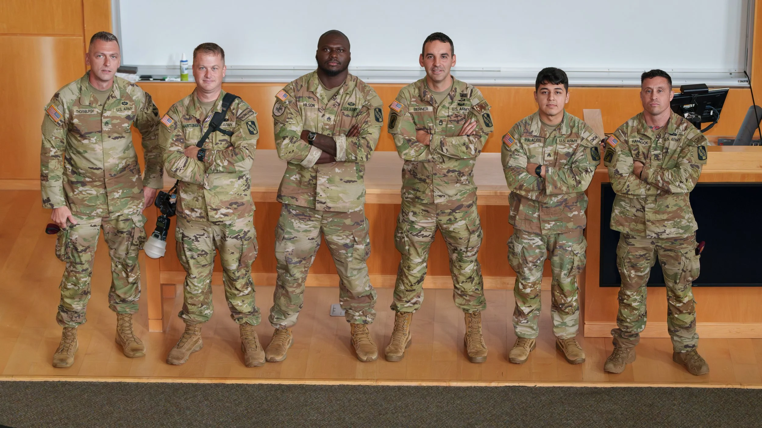 Seven soldiers in camouflage military uniforms standing in a row in a classroom, with a whiteboard behind them.