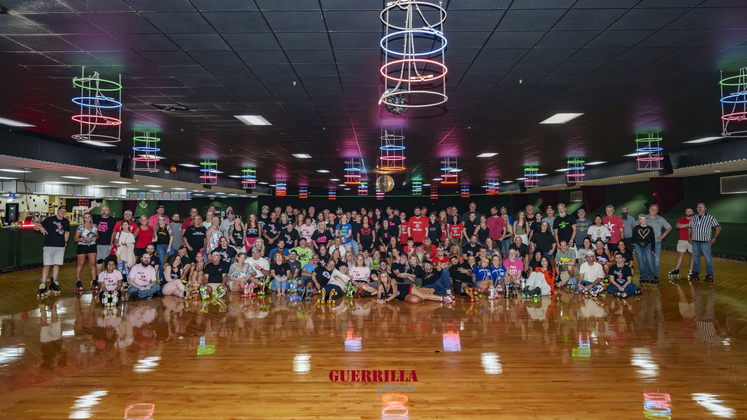 A large group of people gathered in a roller skating rink with colorful neon lights and a shiny wooden floor.