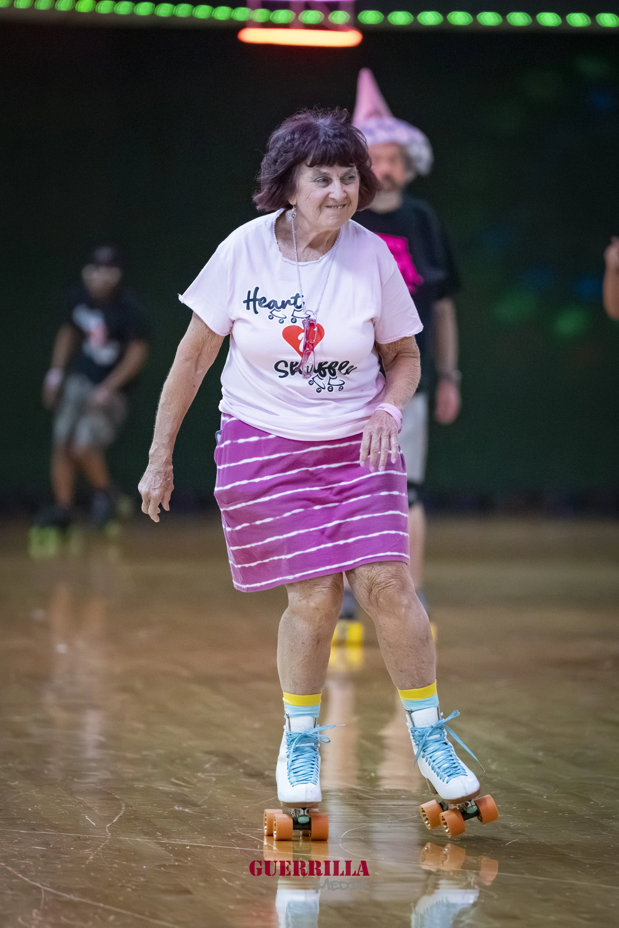 An elderly woman roller skates indoors, wearing a pink skirt, a white t-shirt with a heart and text, and blue roller skates with orange wheels. She is smiling and appears to be enjoying herself.