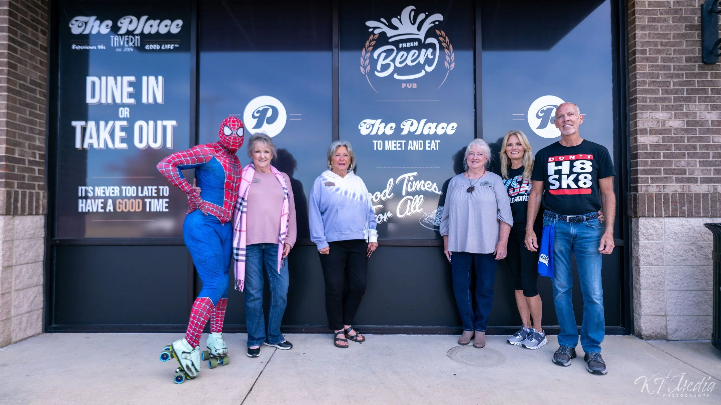 Group of six people, including a person in a Spider-Man costume, standing outside a glass storefront with signs for a pub named 'The Place' and a message about dine-in and take-out options.