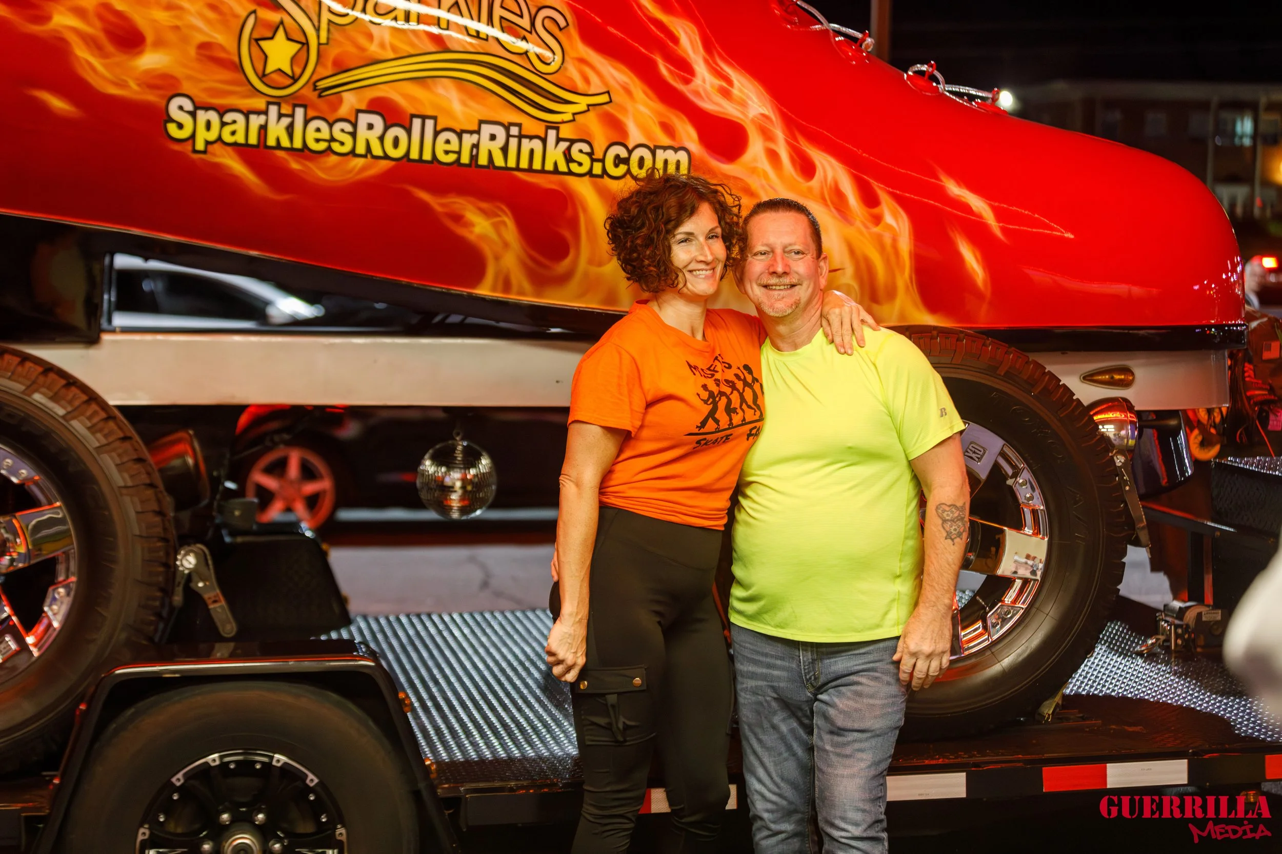 A man and woman smiling posing together in front of a colorful roller coaster vehicle with flames and the website 'SparklesRollerRinks.com' on it, at night.