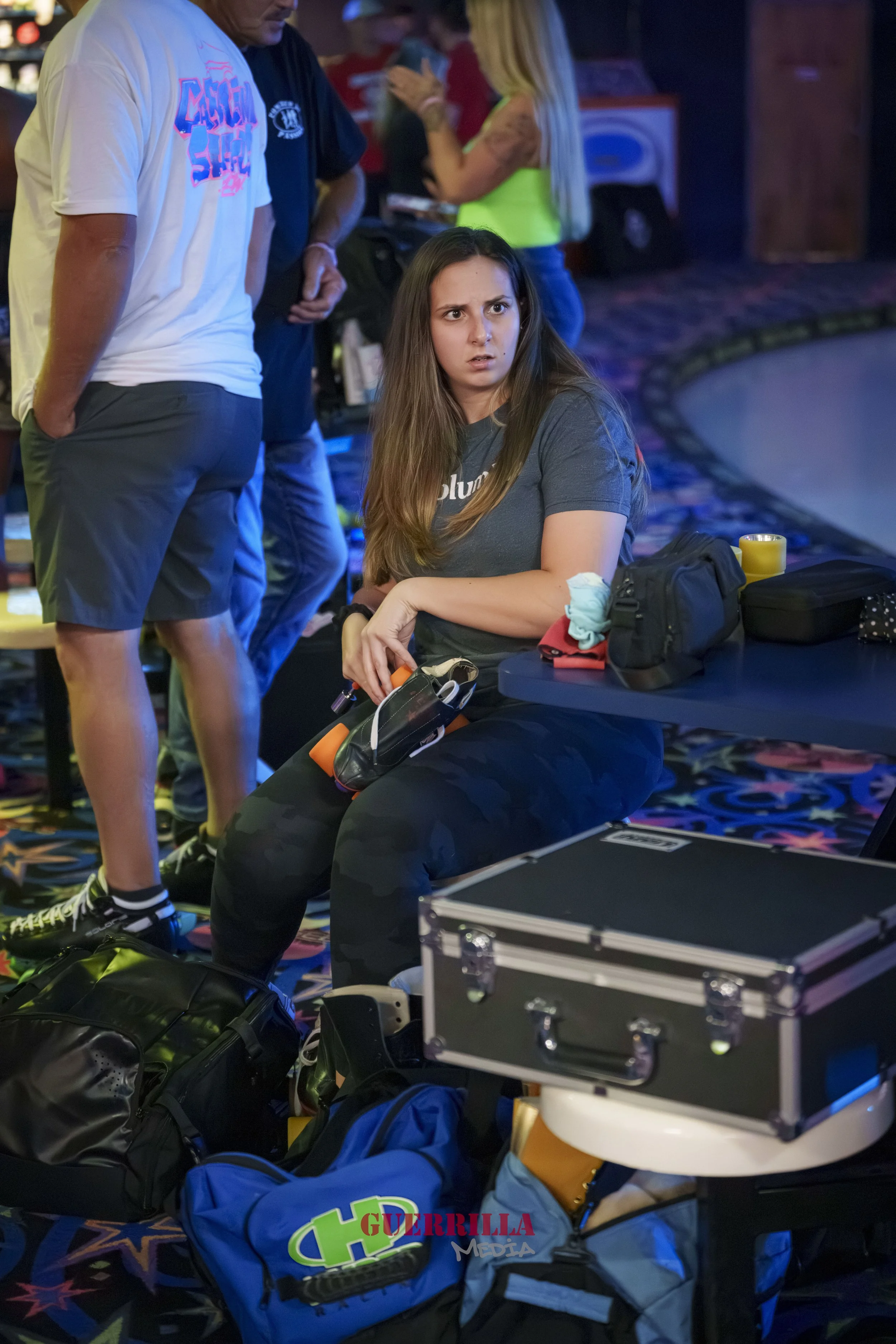 A young woman sitting on a bench at an indoor event, looking confused or surprised, surrounded by backpacks and personal items, with a group of people in the background.