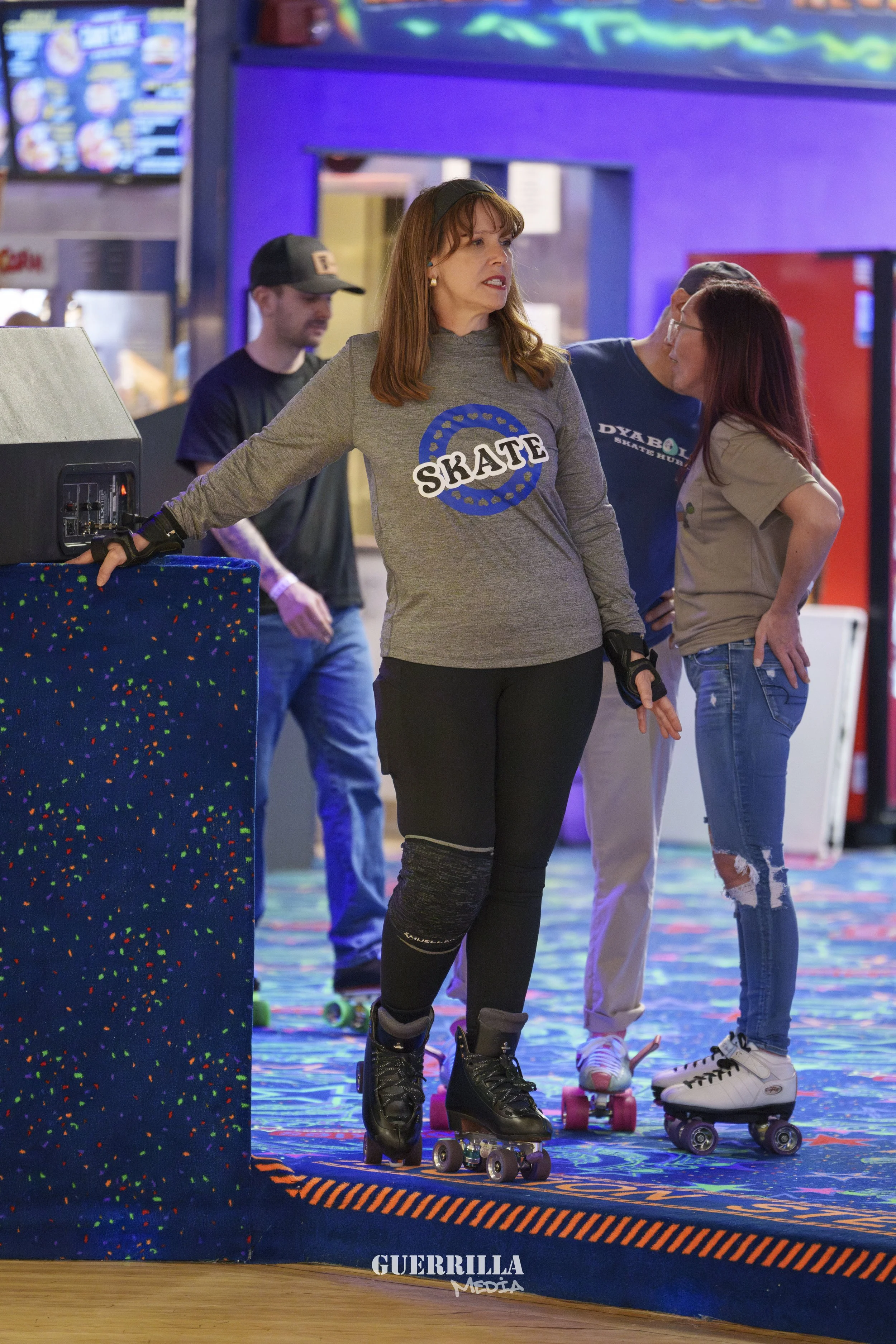 A woman with brown hair wearing a gray long-sleeve shirt with a skate logo, black pants, and roller skates, standing on a roller rink. She appears to be speaking or making a gesture, with two other women and a man in the background, all on roller ska