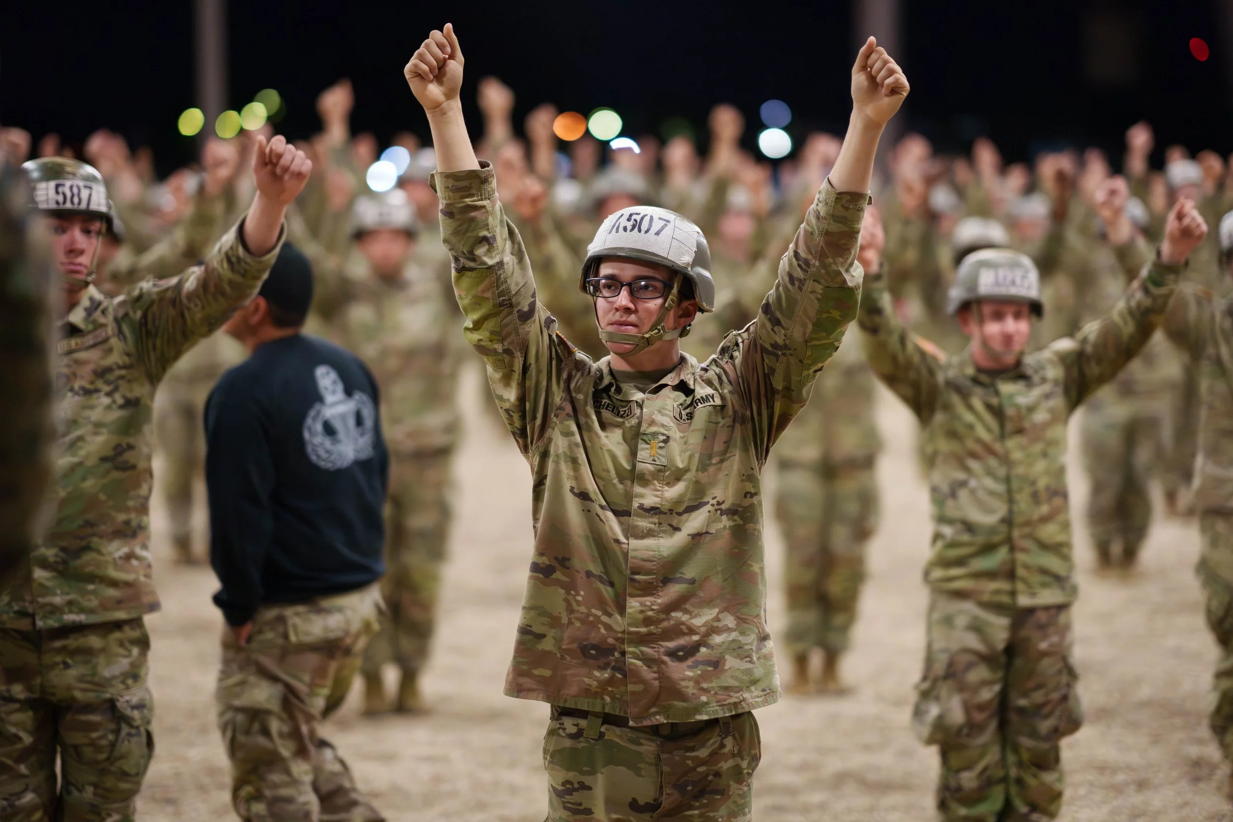 A crowd of soldiers in camouflage uniforms and helmets standing in formation at night, with one soldier in front raising his arms.