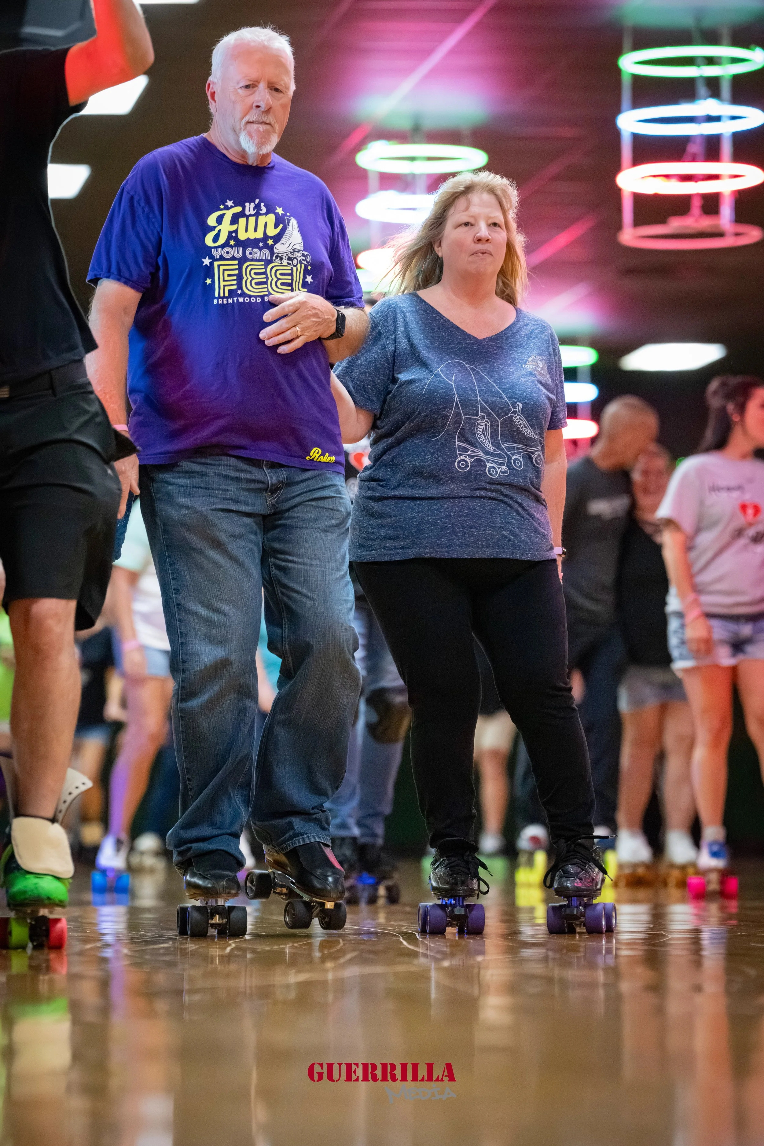 A group of people roller skating indoors, with colorful neon lights overhead. Two people are holding hands, one man with a beard wearing a purple T-shirt and another woman with blonde hair wearing a blue T-shirt featuring a roller skate design.