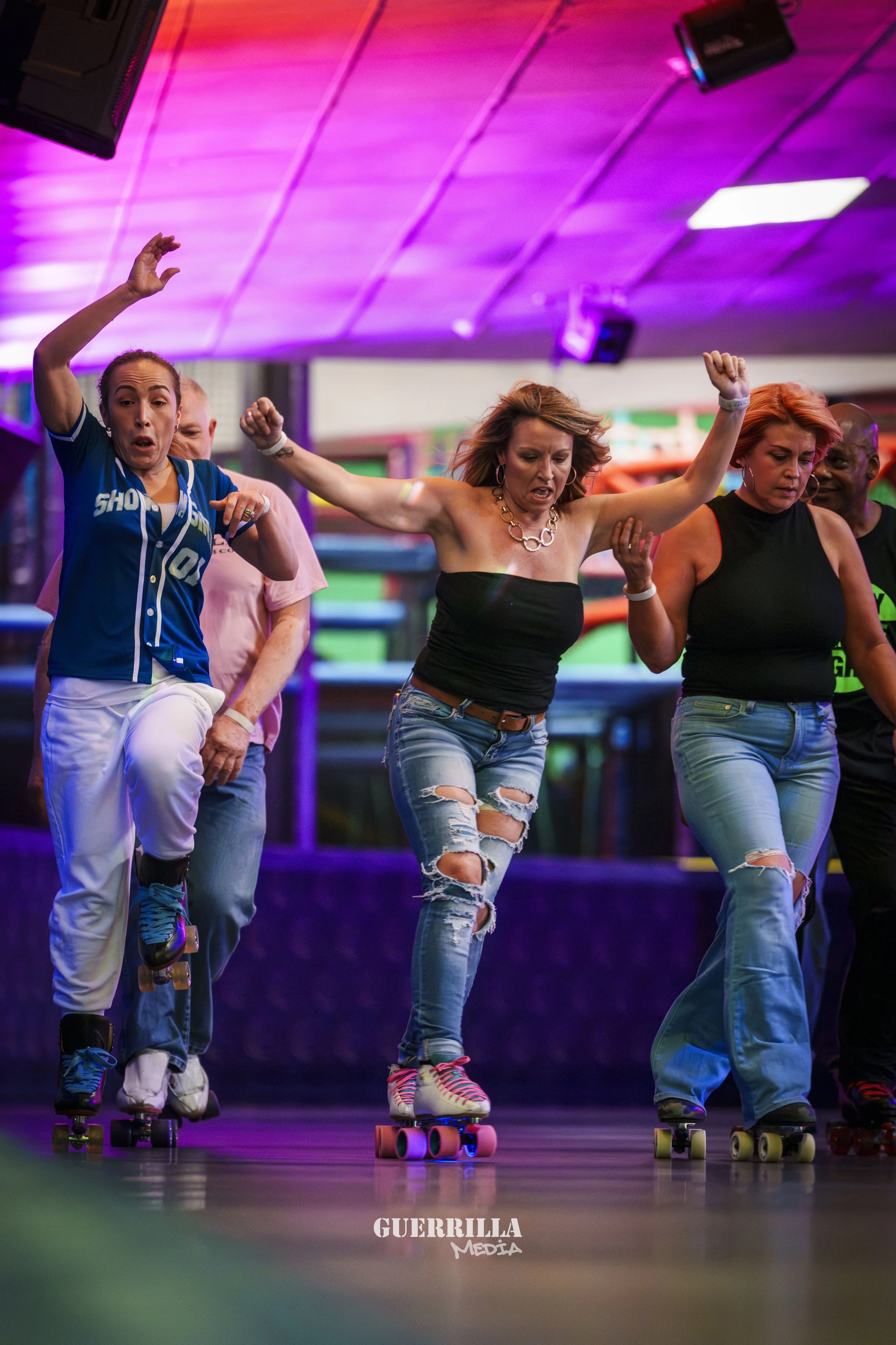 A group of women roller skating indoors, dressed casually, with some raising their arms and others concentrating on skating, under colorful lighting.
