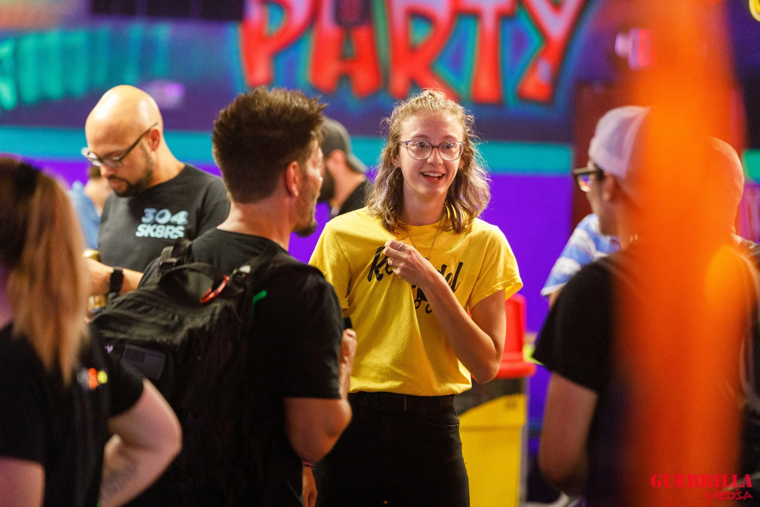 A group of people at a colorful party, with a woman in a yellow shirt talking to others. The background has a vibrant, rainbow-colored 'PARTY' sign.