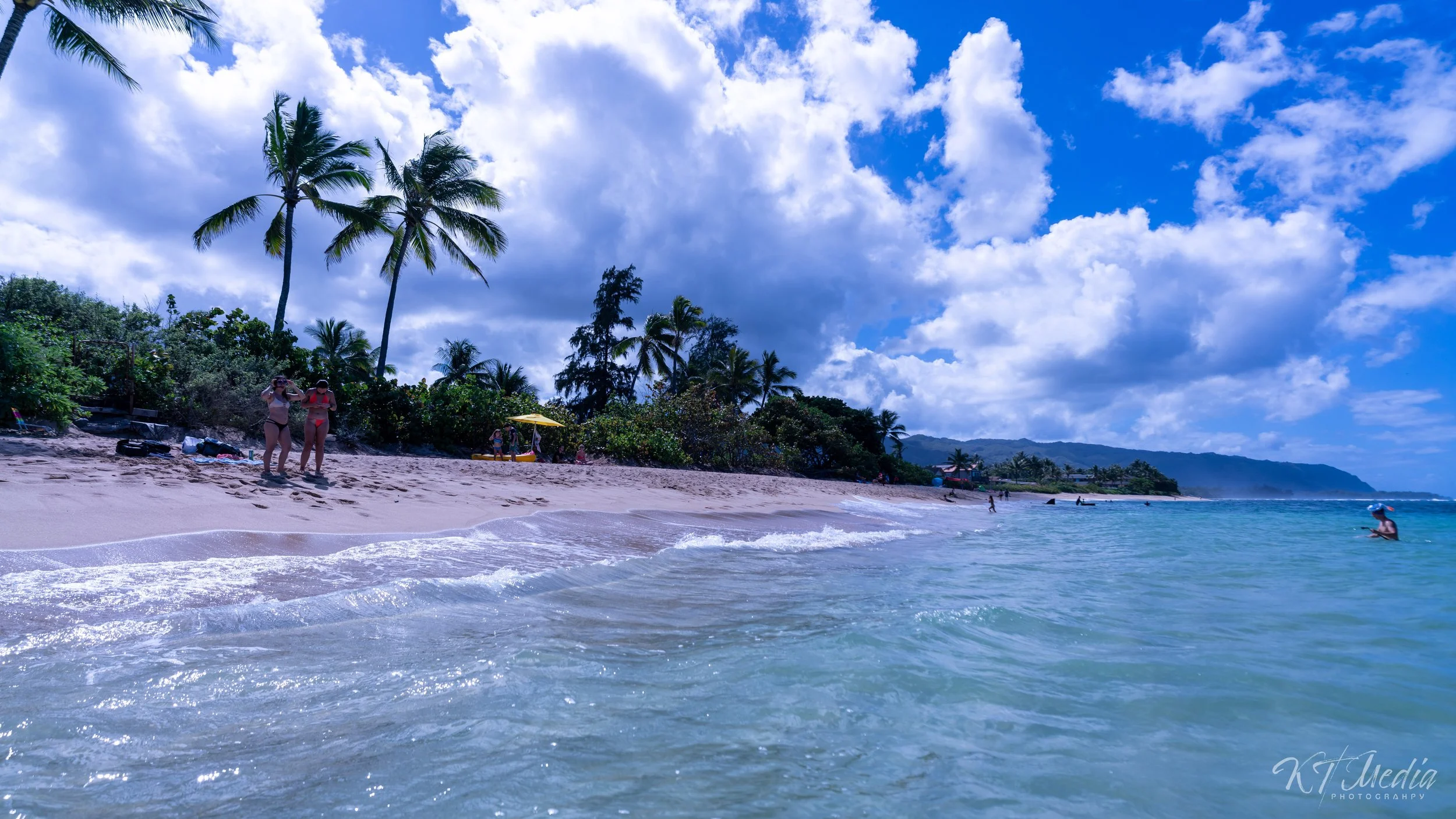 People relaxing and enjoying the beach with palm trees, clouds, and mountains in the background.
