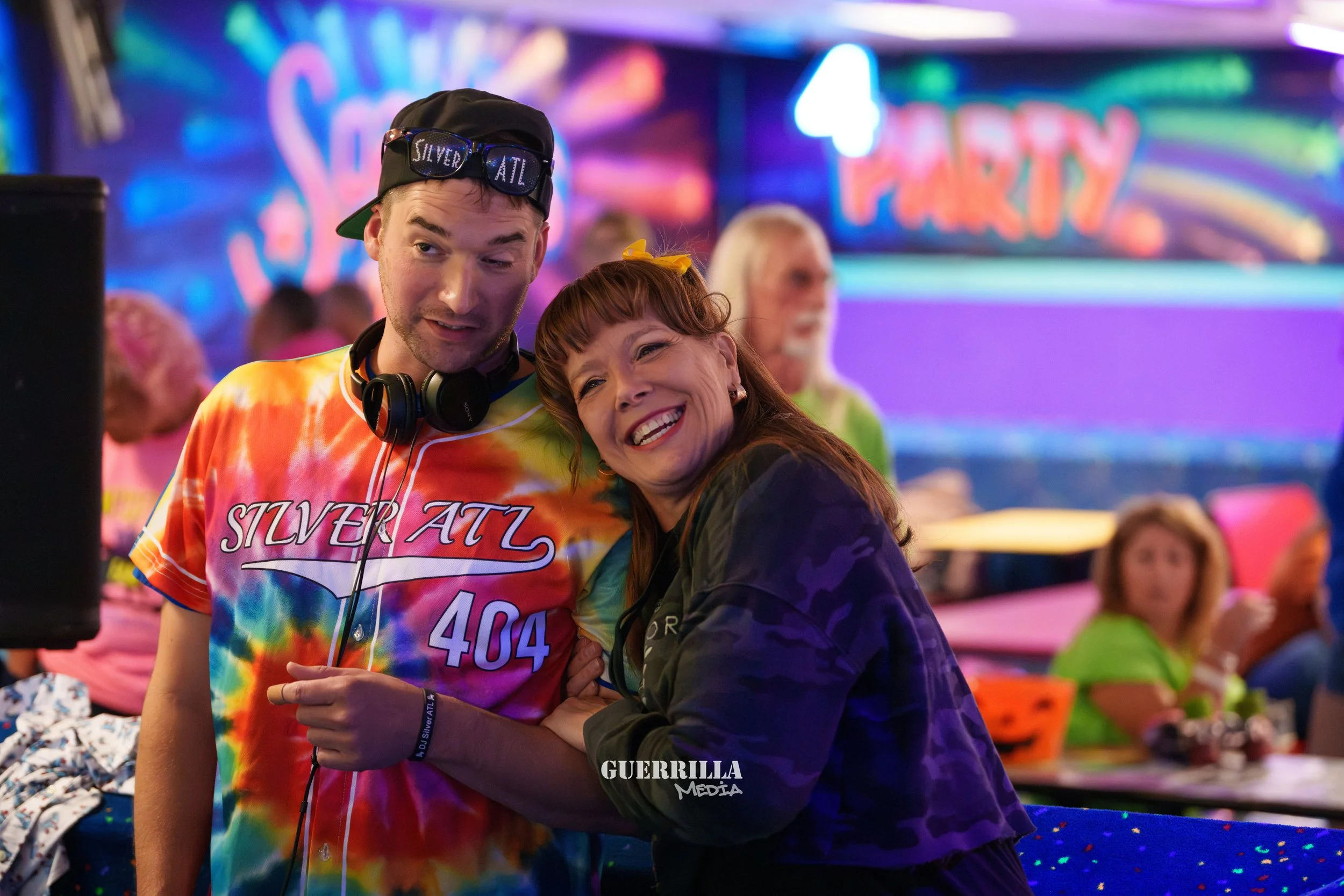 A man and woman smiling and posing together at a party with colorful neon signs in the background, decorations, and other guests.