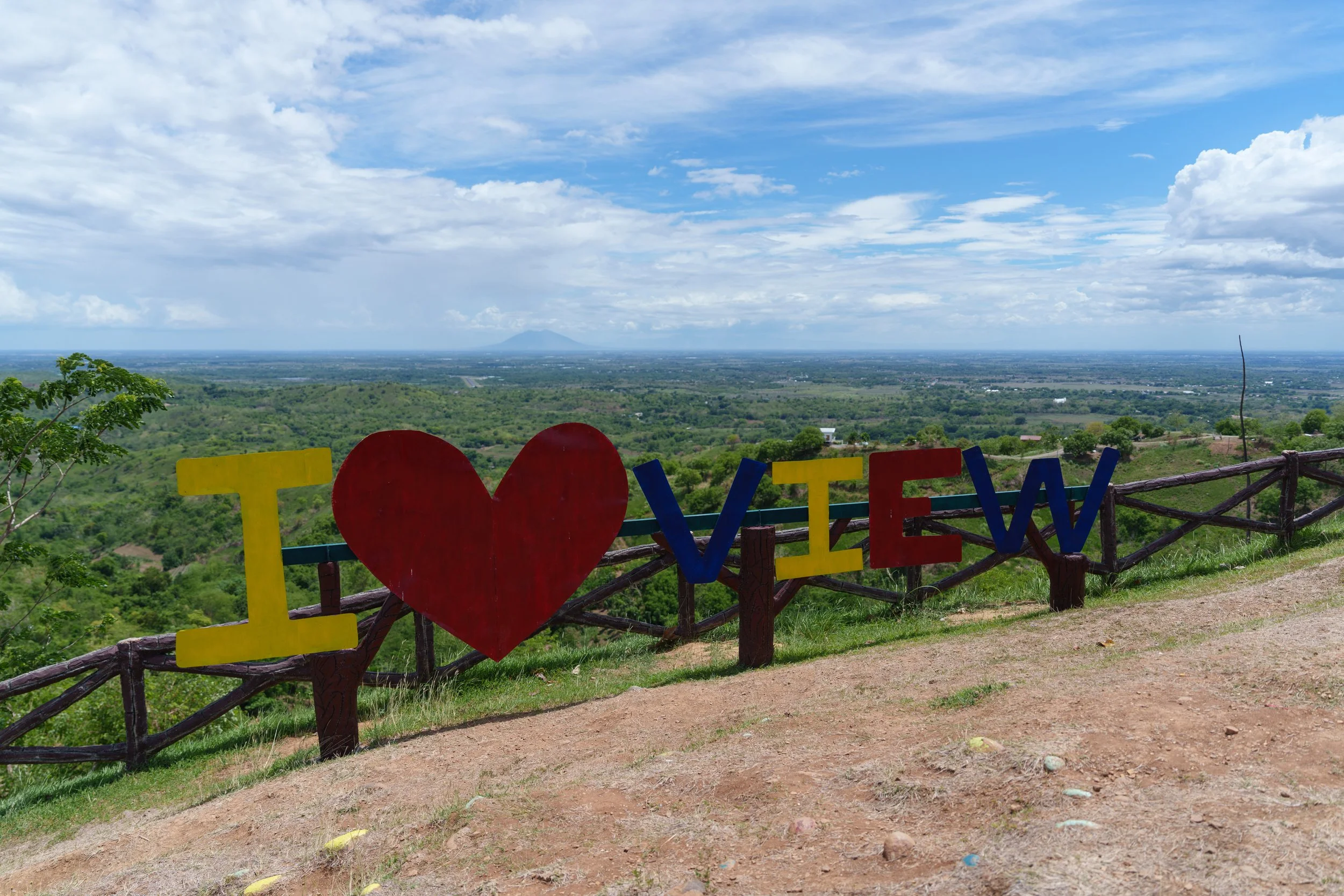 Colorful sign reading 'I love VIEW' with a heart symbol replacing the word 'love,' set against a scenic landscape with green fields, a blue sky with clouds, and a mountain in the distance.