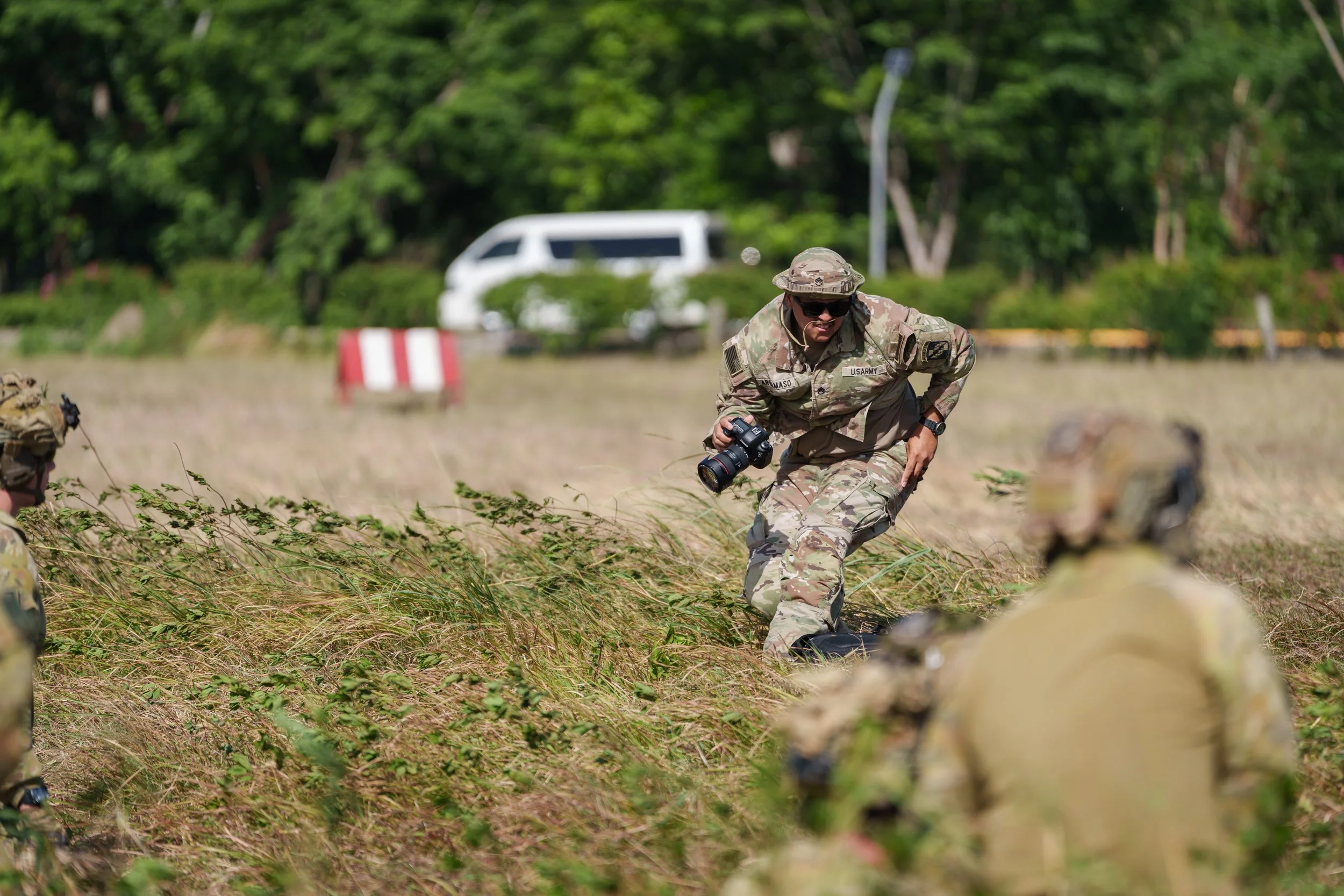 Military personnel in camouflage uniform during field training, with one person holding a camera, in a grassy field with trees and a white vehicle in the background.