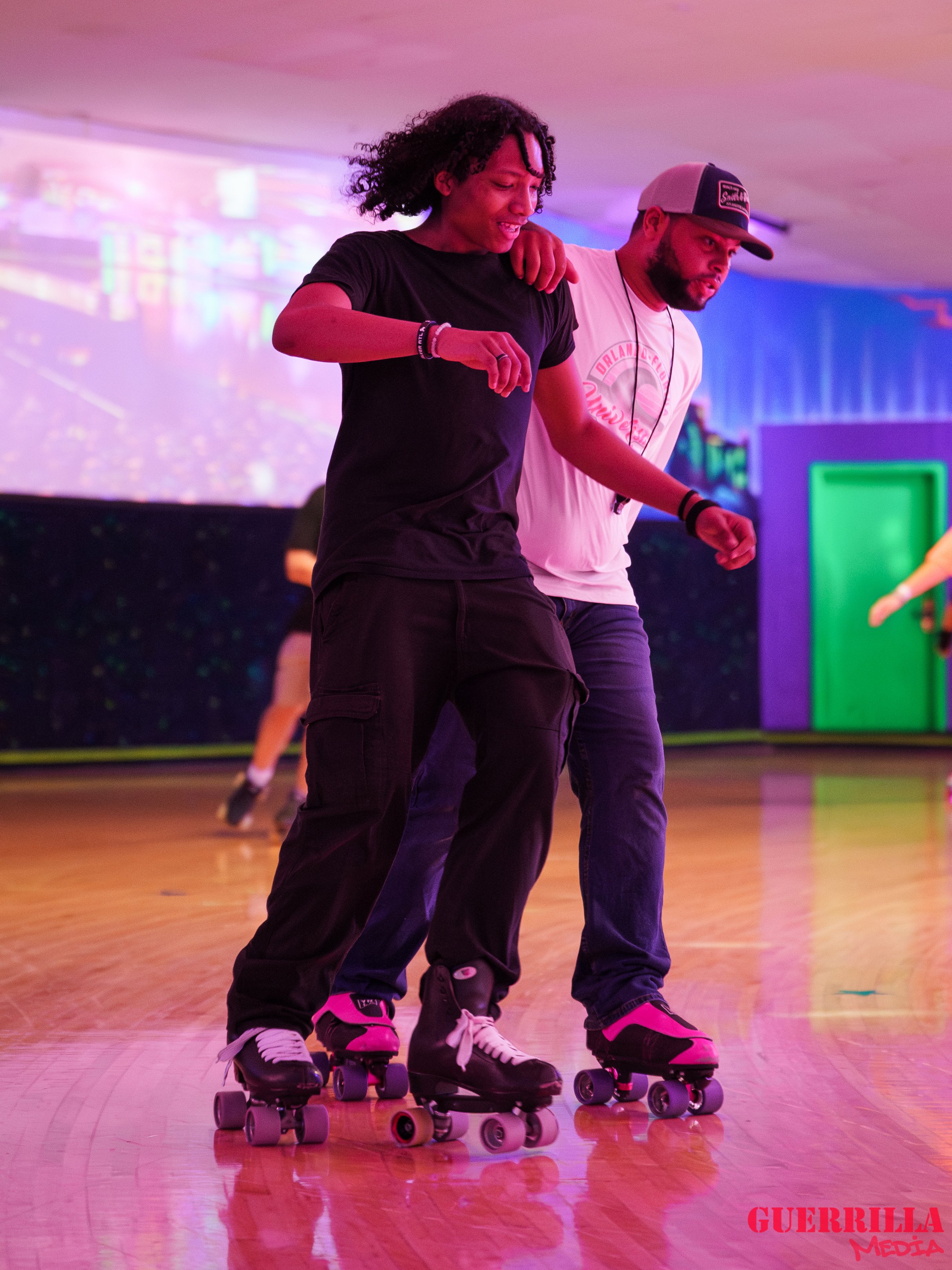 Two people roller skating together indoors under colorful lights, with one person helping the other maintain balance.