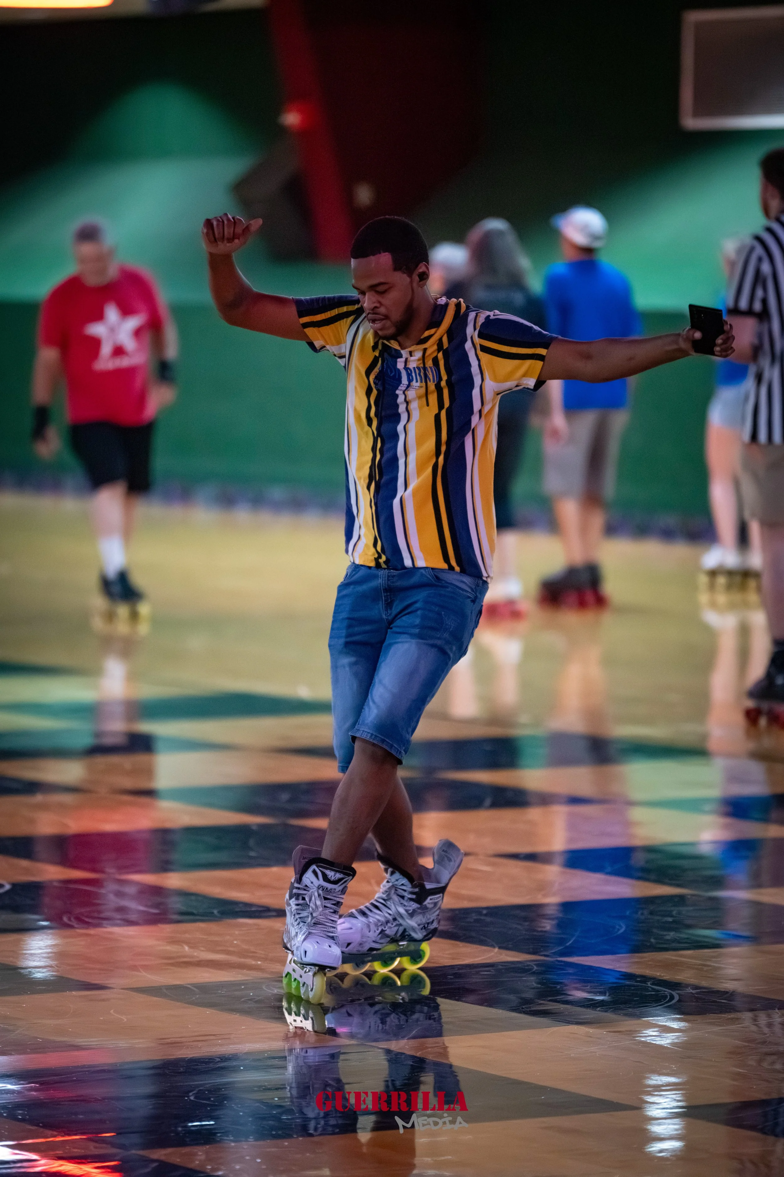 A man roller skating indoors on a checkered floor, wearing a striped yellow, black, and blue shirt, denim shorts, and white roller skates, with others in the background and the word 'GUERRILLA' at the bottom of the image.