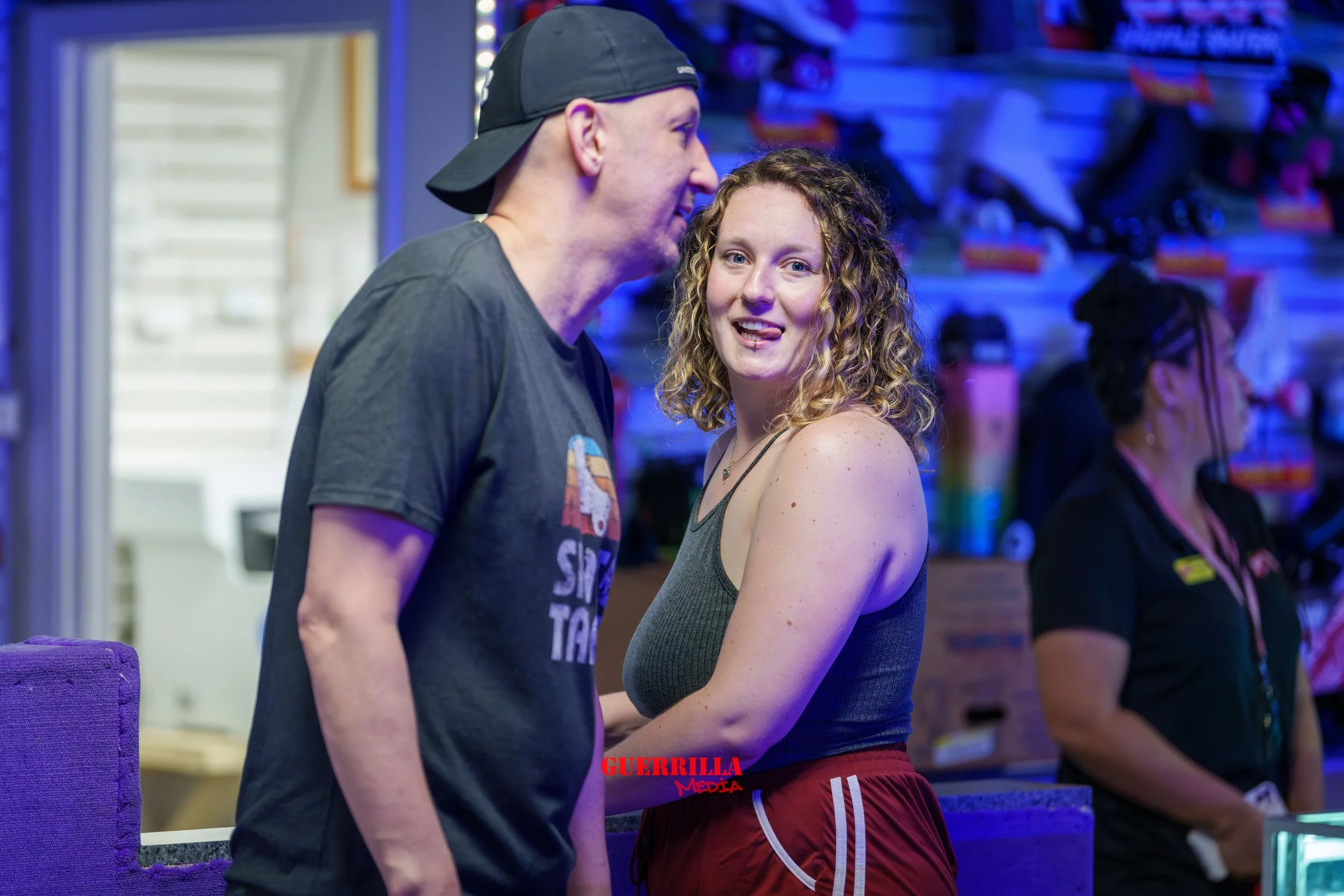 A young woman with curly hair and a tongue piercing in a casual, sleeveless top and red shorts, smiling and engaging with a man wearing a black cap backward and a dark T-shirt, in a colorful retail or arcade environment.