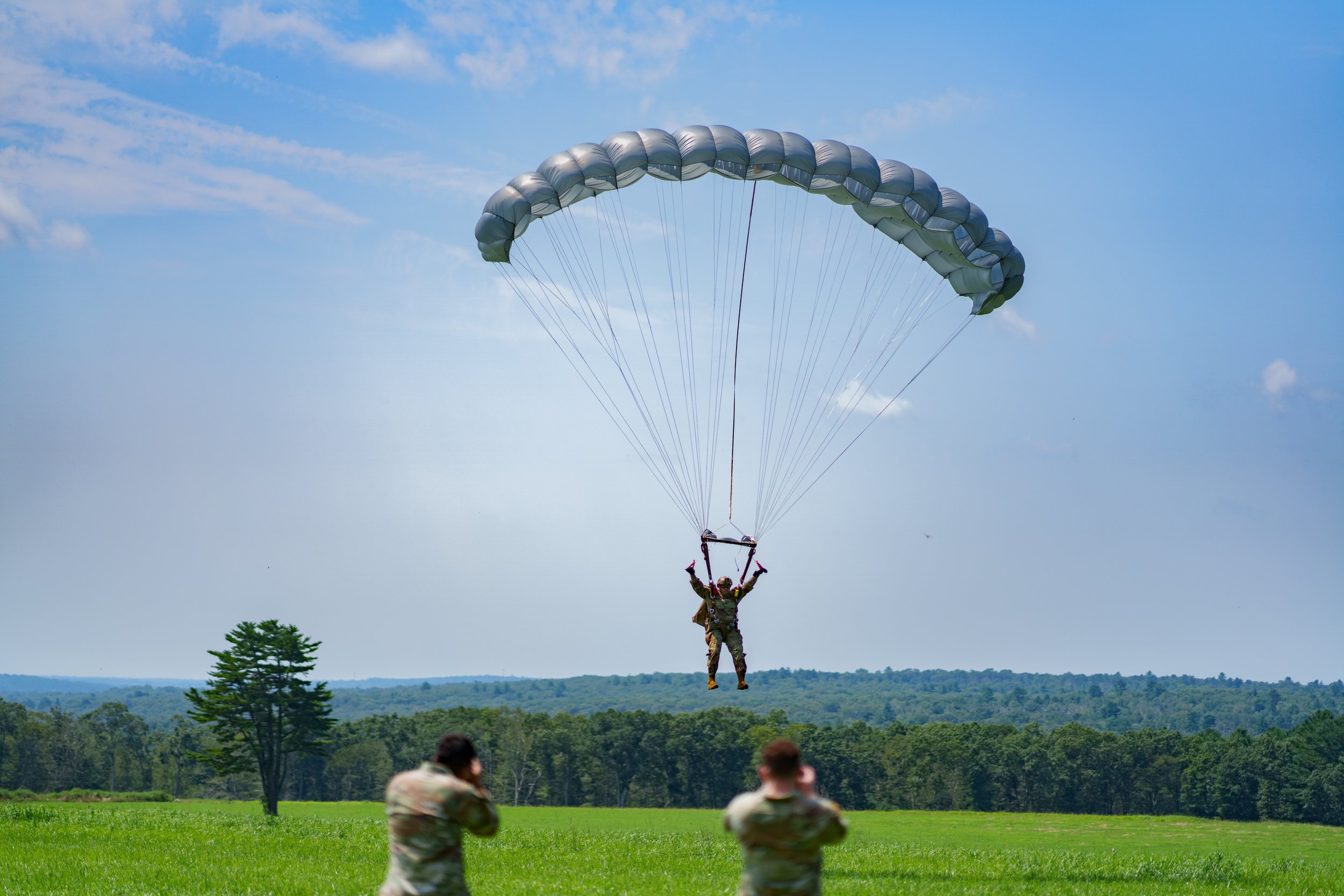 A soldier parachuting down with a silver parachute while two soldiers on the ground take photos or videos in a green field with trees in the background.