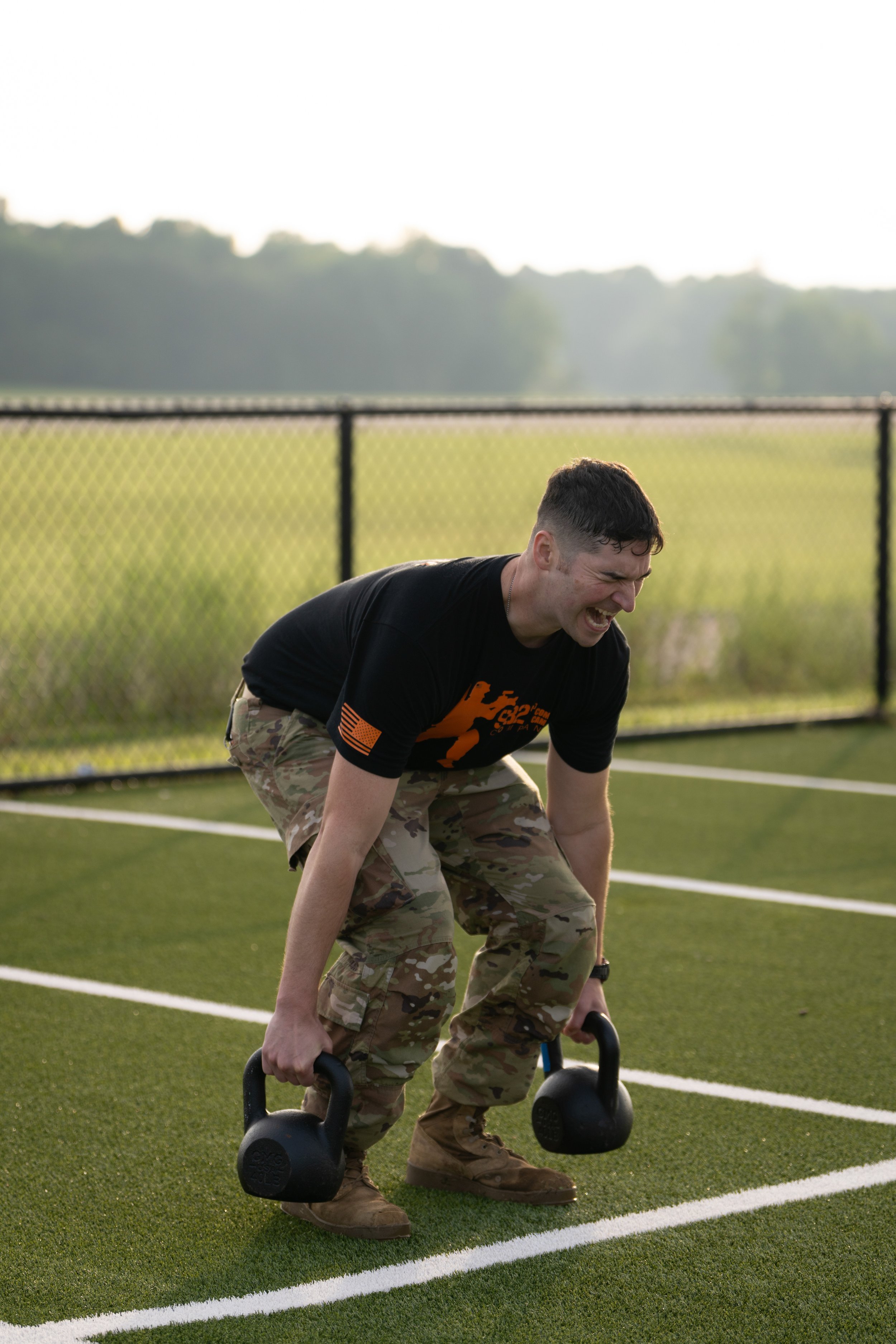 A man in camouflage pants and a black T-shirt doing a workout with kettlebells on a sports field.