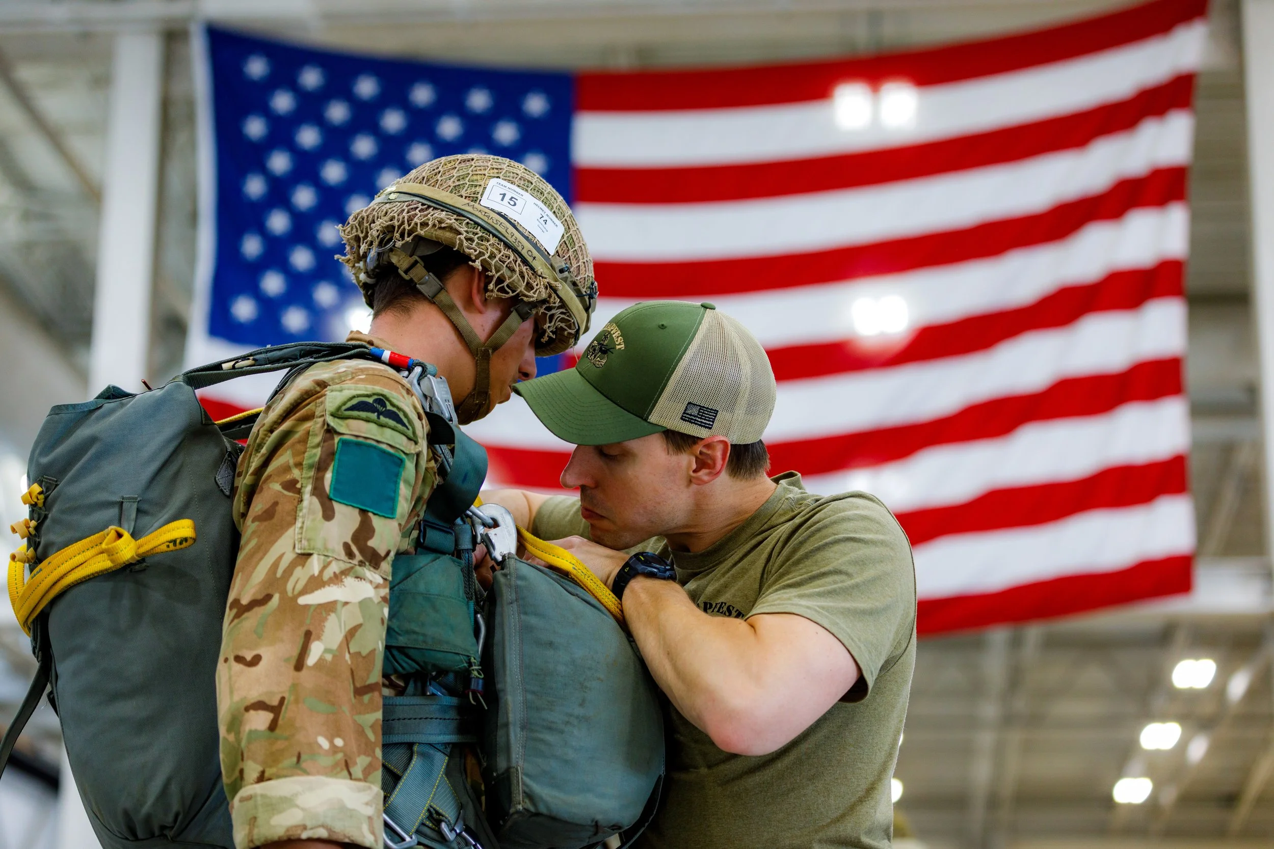 A soldier in camouflage uniform and helmet is being assisted by a man in a green t-shirt and cap, with a large American flag hanging in the background.