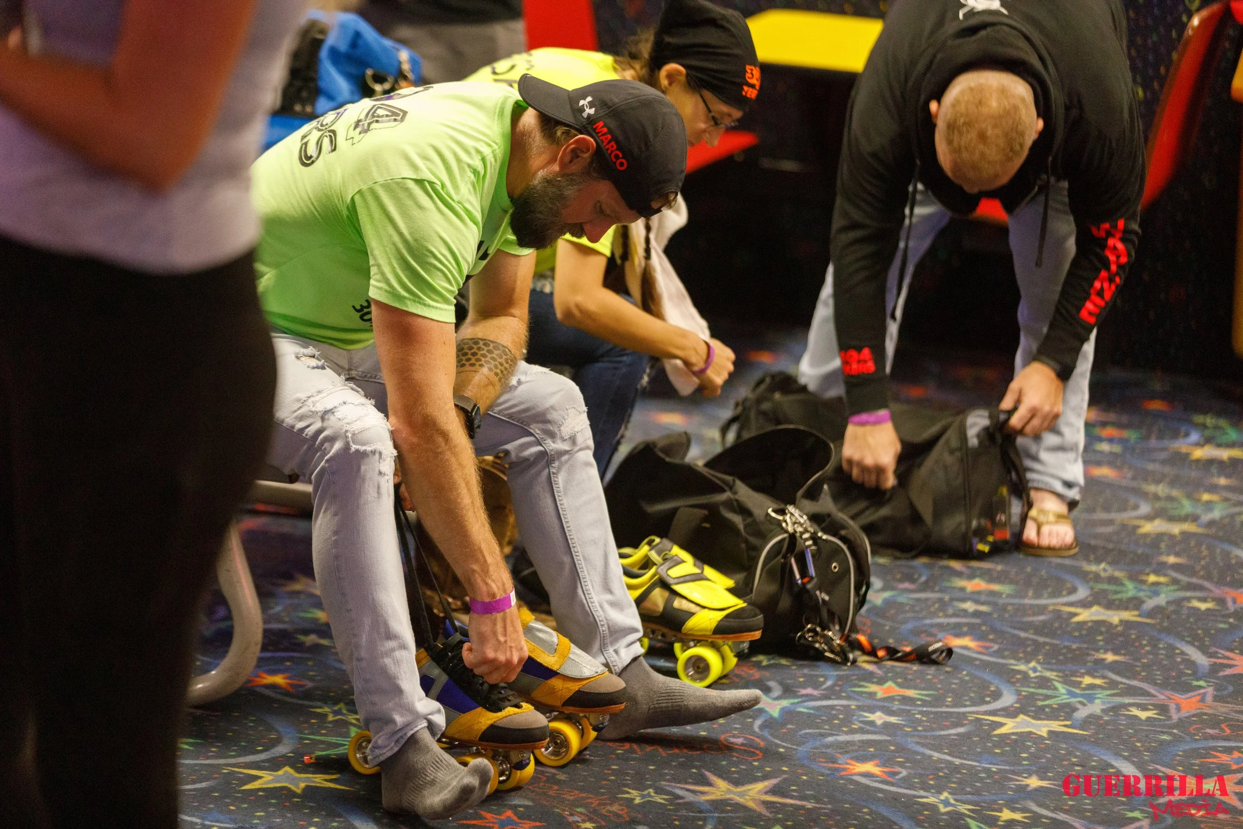 Two men sitting on the floor putting on roller skates with their gear on their laps. A woman and another man are also beside them, preparing their skates. They are on a colorful, starry-patterned carpeted surface in an indoor skating rink or arena.