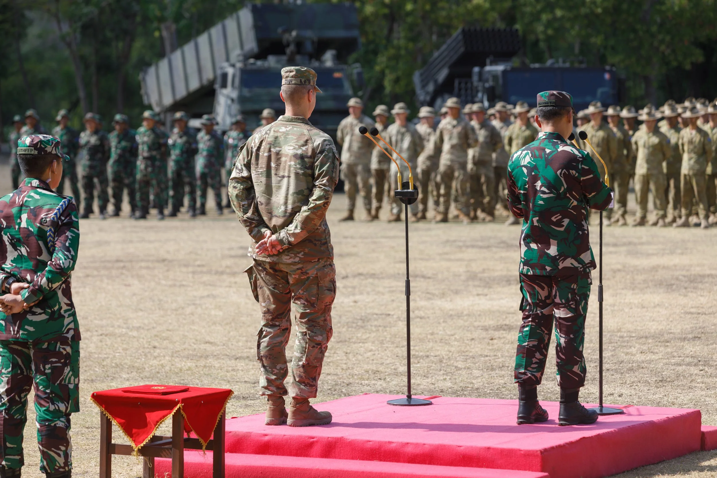 Military officers and soldiers in camouflage uniforms standing at attention during a ceremony outside, with leaf trees in the background.