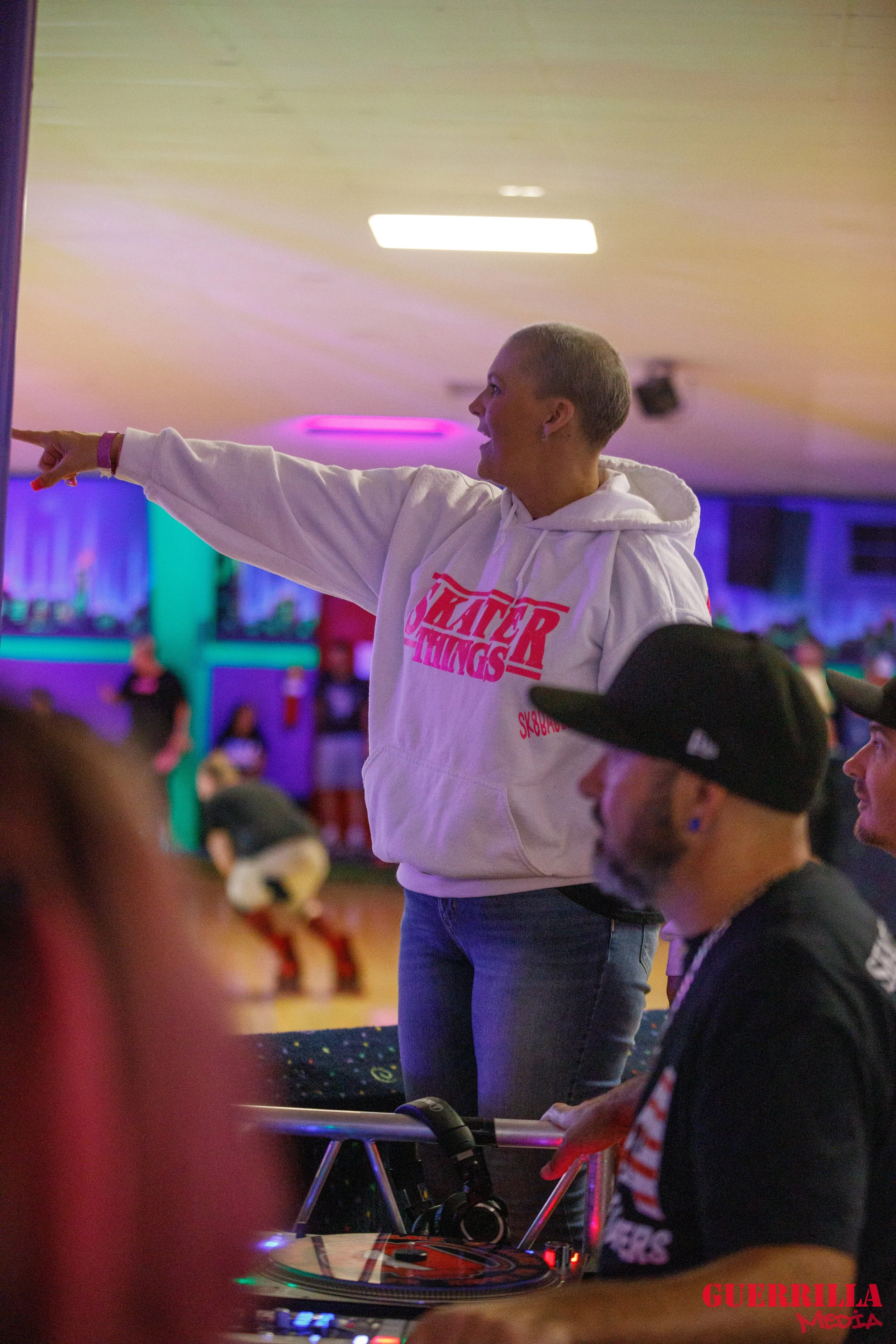 A woman with short gray hair wearing a white hoodie with red text pointing and talking to a group. In the foreground, a man wearing a black cap and striped shirt is looking in the same direction, with a DJ turntable and headphones nearby. The backgro