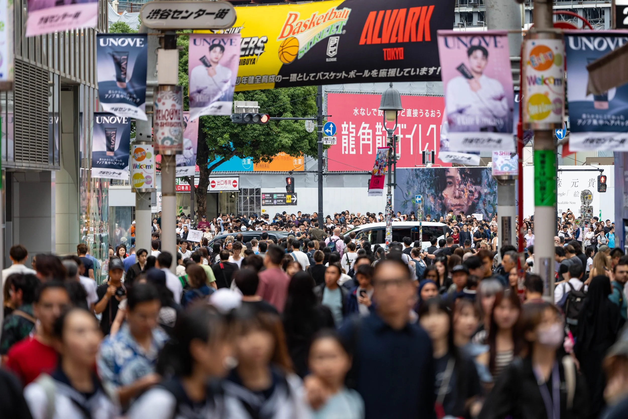 A crowded city street filled with pedestrians and various colorful signs and billboards, including advertisements in Japanese and English.