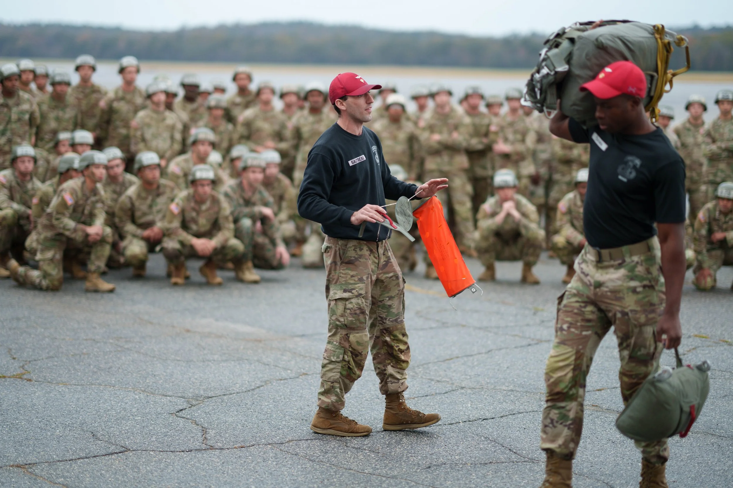 Military personnel in camouflage uniforms participating in a briefing outdoors, with a water body in the background.