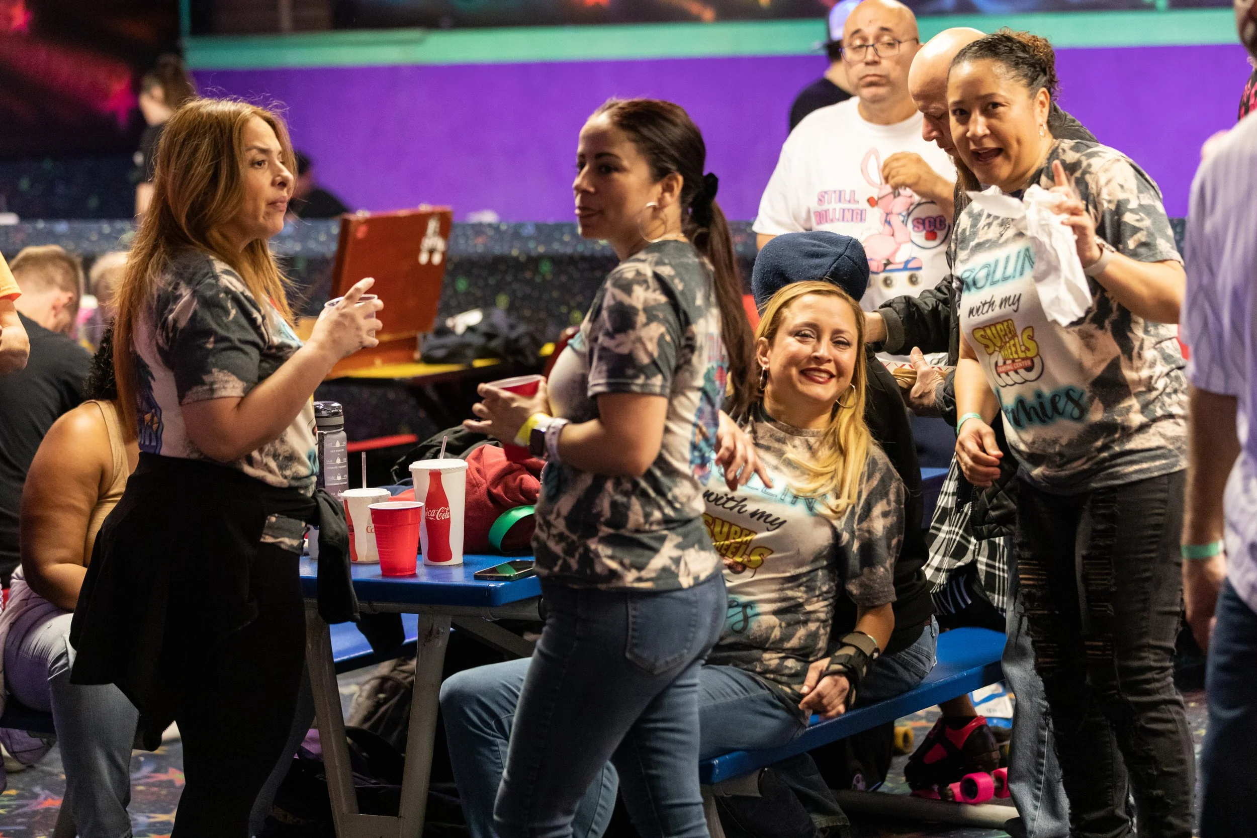 Group of women and a girl gathering at a roller skating rink, some wearing matching T-shirts, with drinks and skate gear on the table, engaged in conversation.