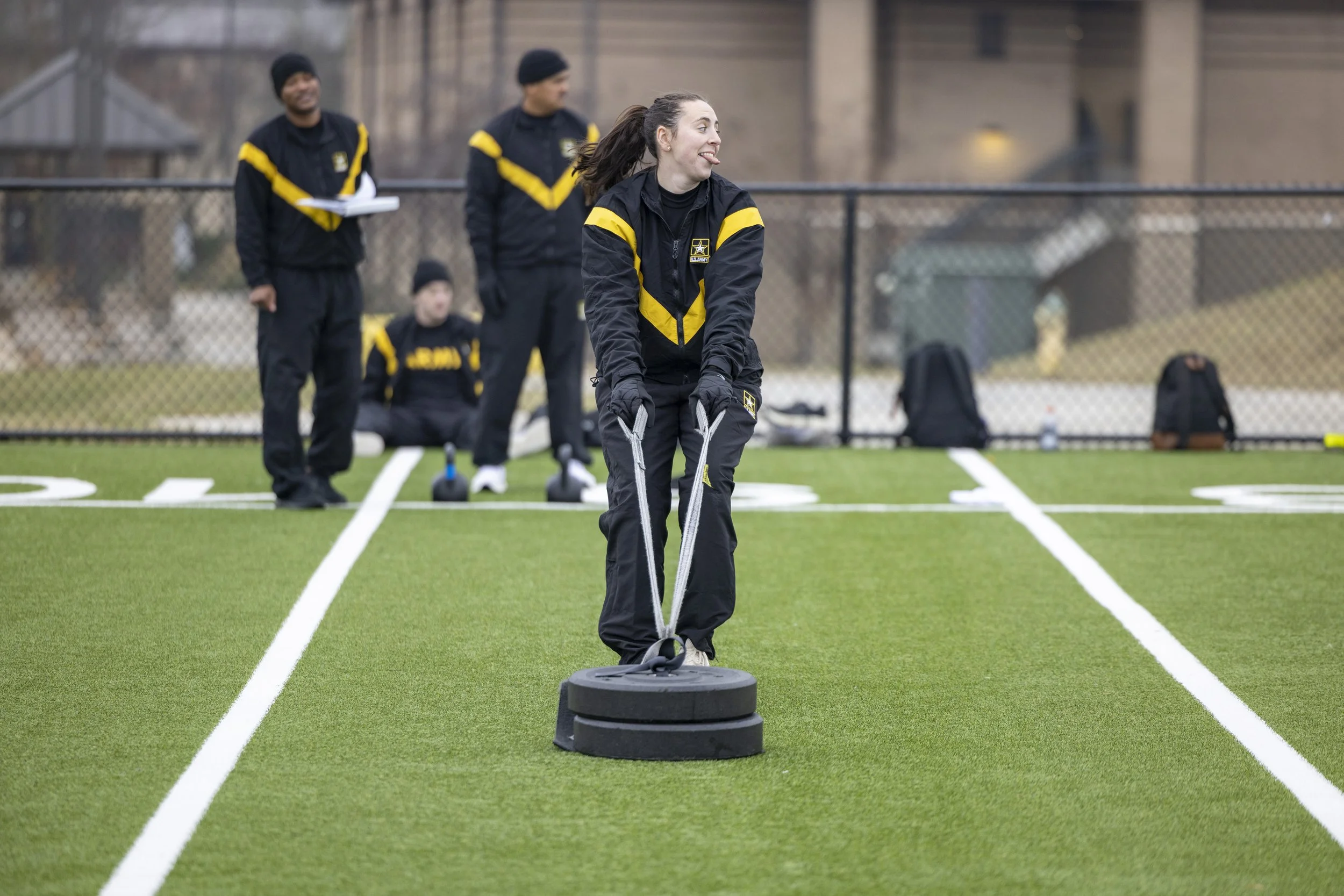 A woman in military-style uniform smoothing a football field with a weighted tool while three men in similar uniforms supervise in the background.