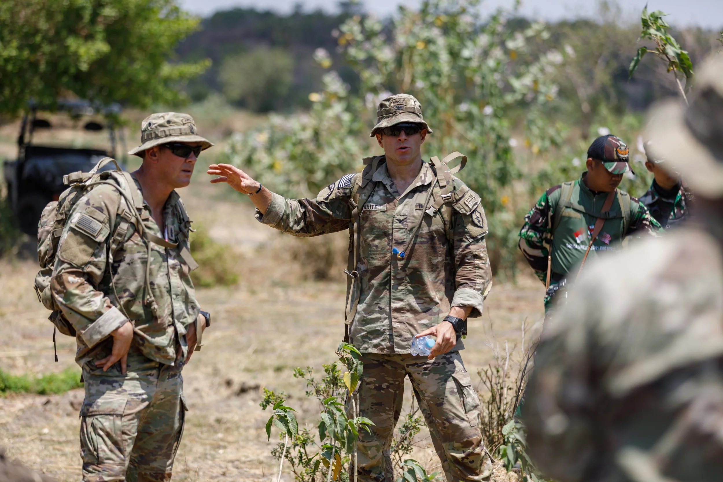 A group of military personnel in camouflage uniforms and backpacks engaged in a discussion outdoors with trees and vegetation in the background.