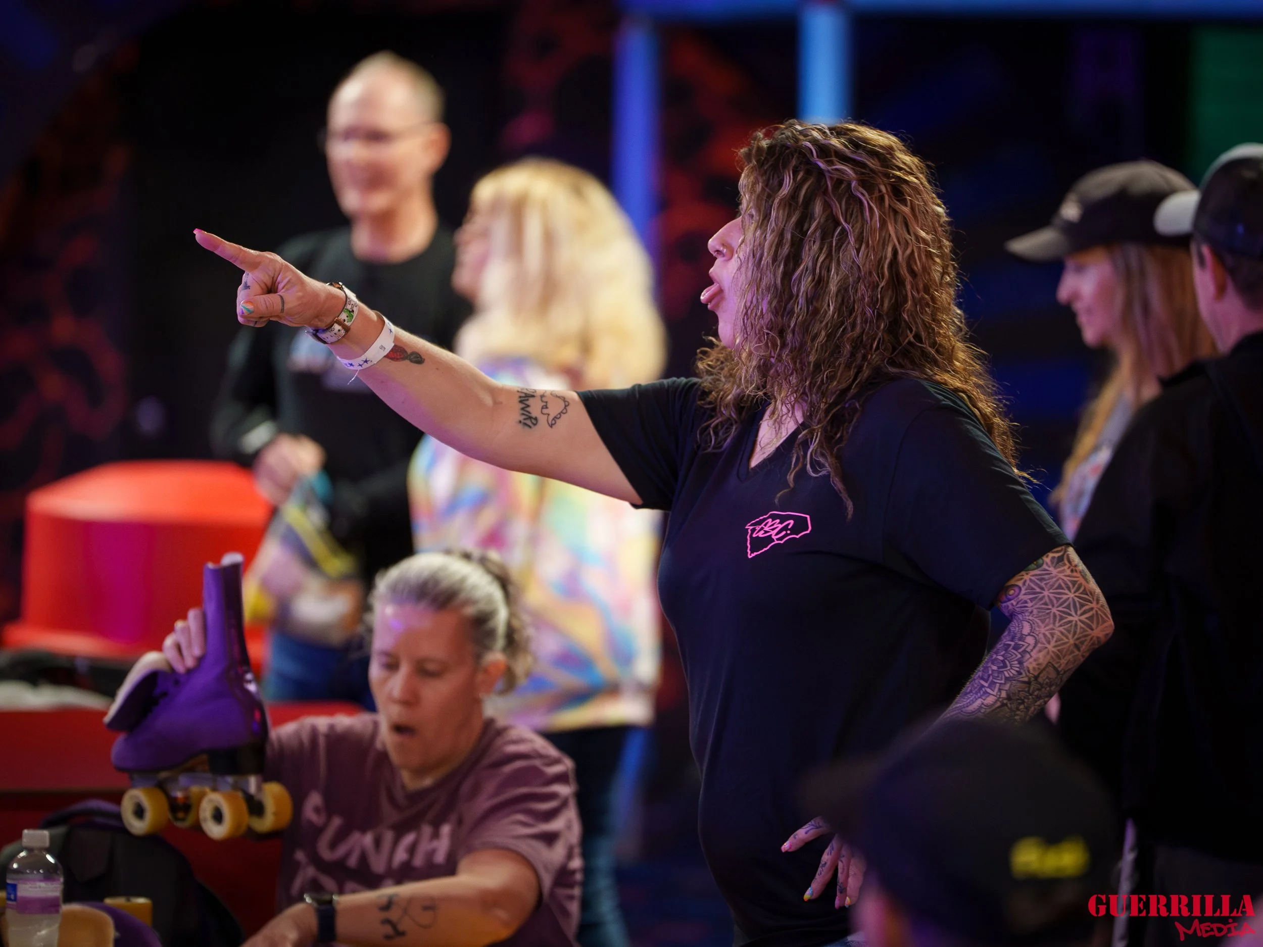 A group of people at a roller skating rink. A woman with curly hair and tattoos on her arm is pointing and engaging with others, while a woman in the foreground wearing a brown t-shirt is sitting with purple roller skates nearby.