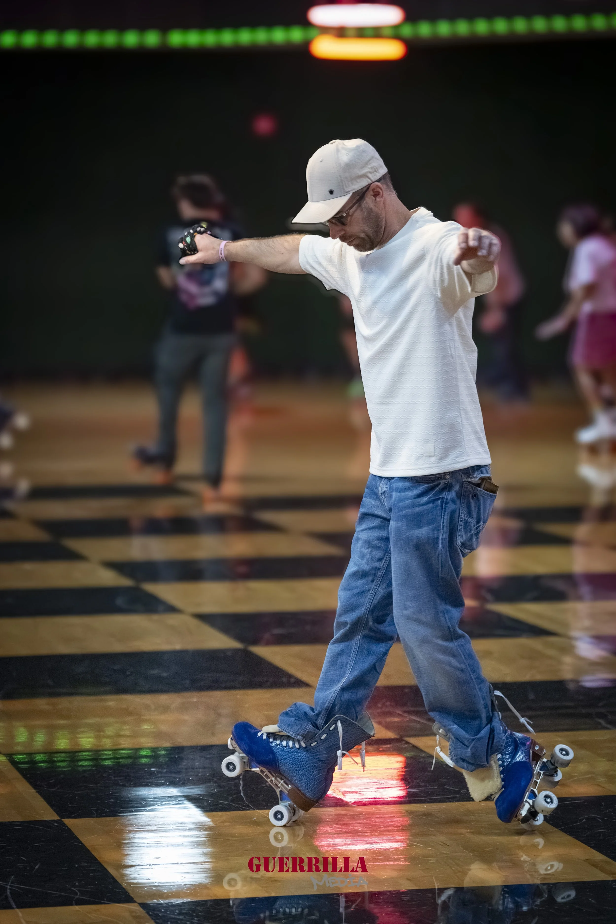 A man in a white t-shirt, jeans, cap, and sunglasses roller skating indoors on a wooden checkered dance floor. People are in the background, slightly out of focus, with colorful lights above. The word 'GUERRILLA' appears in red at the bottom center.