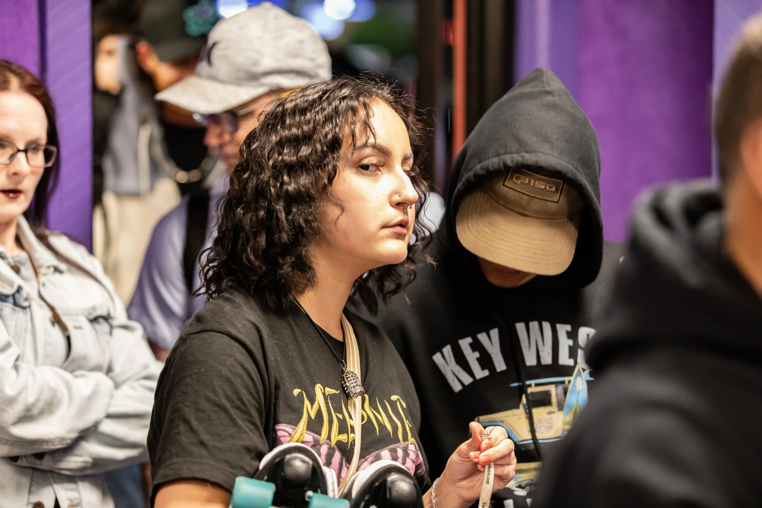 A group of young people at a social gathering, with a woman with curly hair wearing a black Metallica t-shirt and a man in a black hoodie and beige cap leaning in.