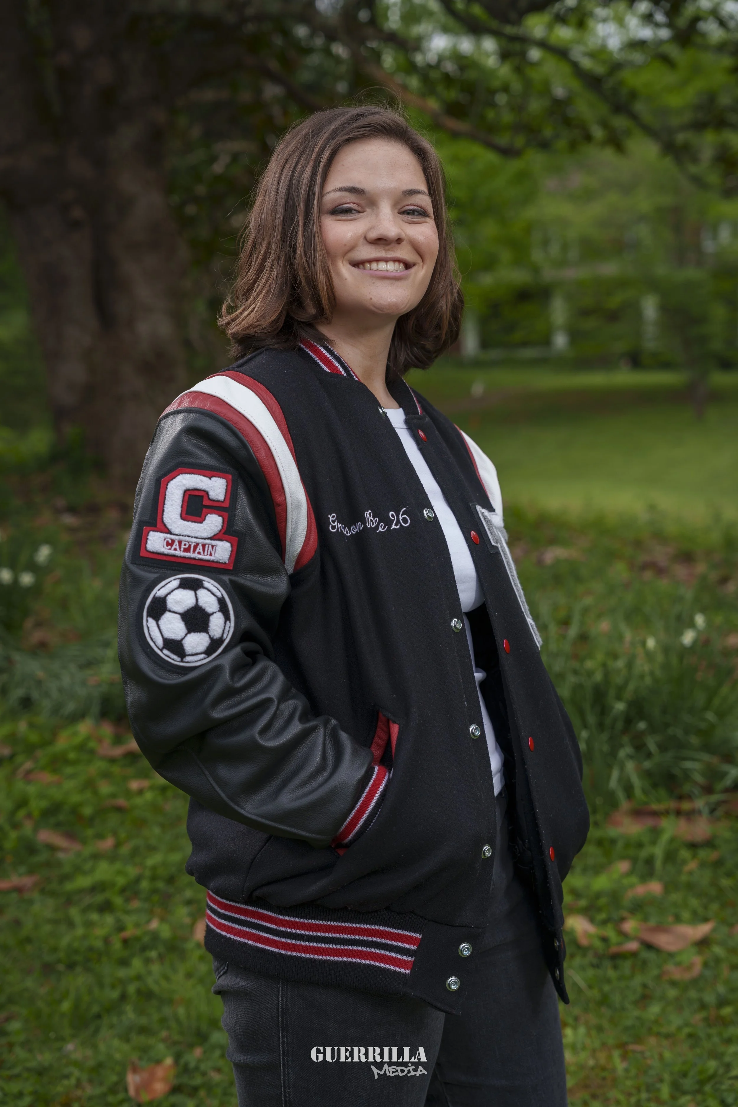 A young woman with shoulder-length brown hair smiling outdoors, wearing a varsity jacket with patches including a soccer ball and a captain's patch, standing in a green park with trees and grass.
